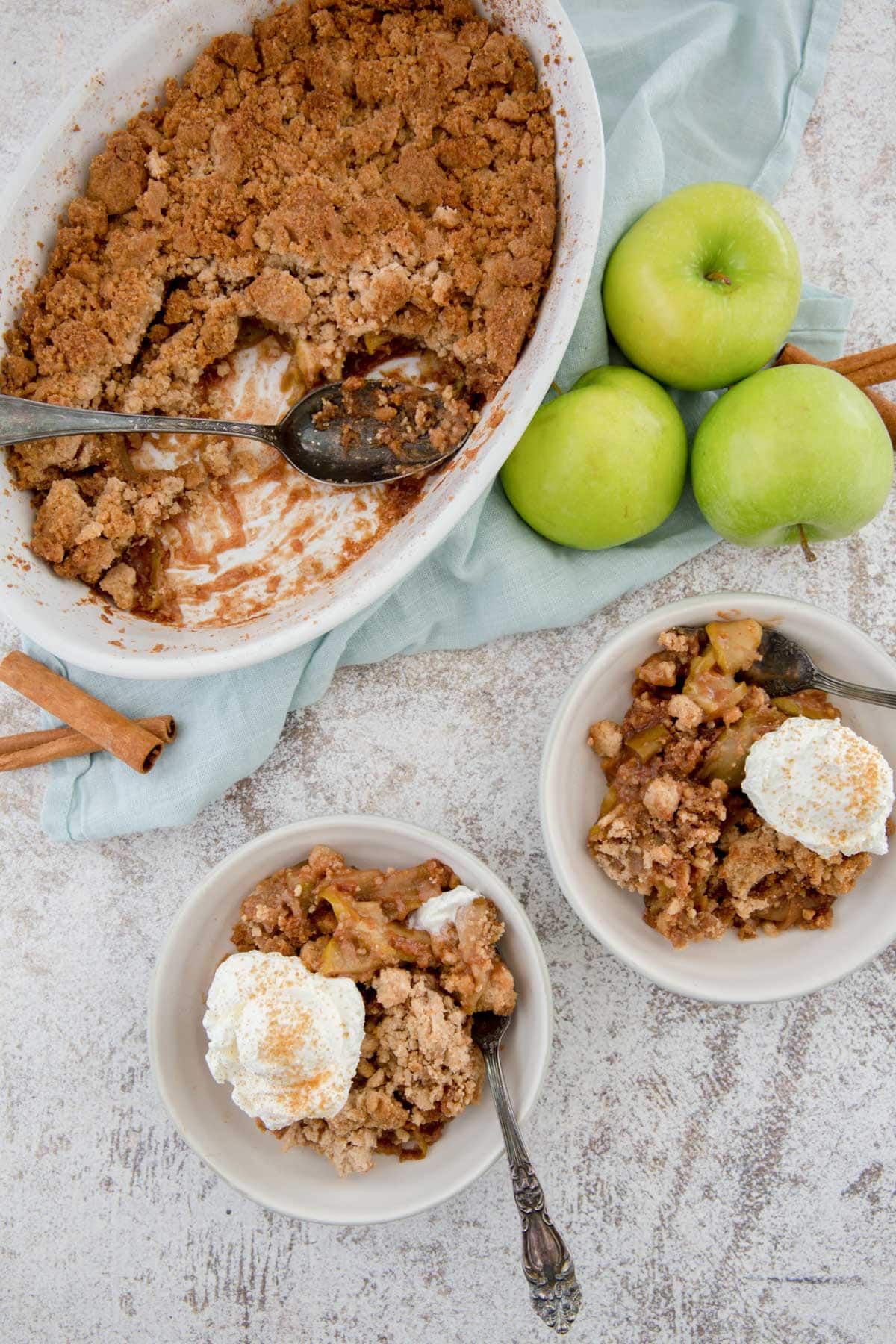 casserole dish with apples and cookie crumble topping, 2 bowls with apple cobbler and whipped cream, green apples, a spoon