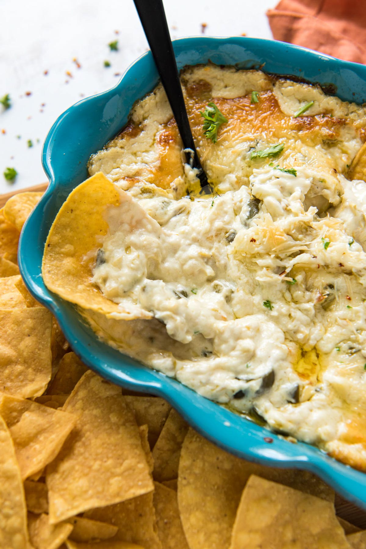 blue dish with jalapeno artichoke dip, tortilla chips, a spoon, an orange napkin.