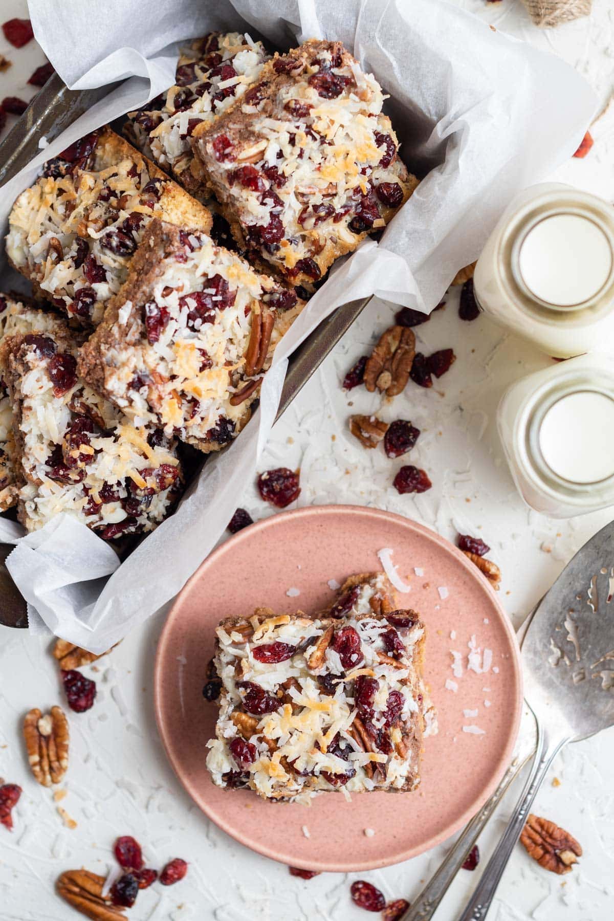 magic bars in a pan, one on a pink plate, 2 jars of milk, scattered chocolate chips, a silver serving spoon