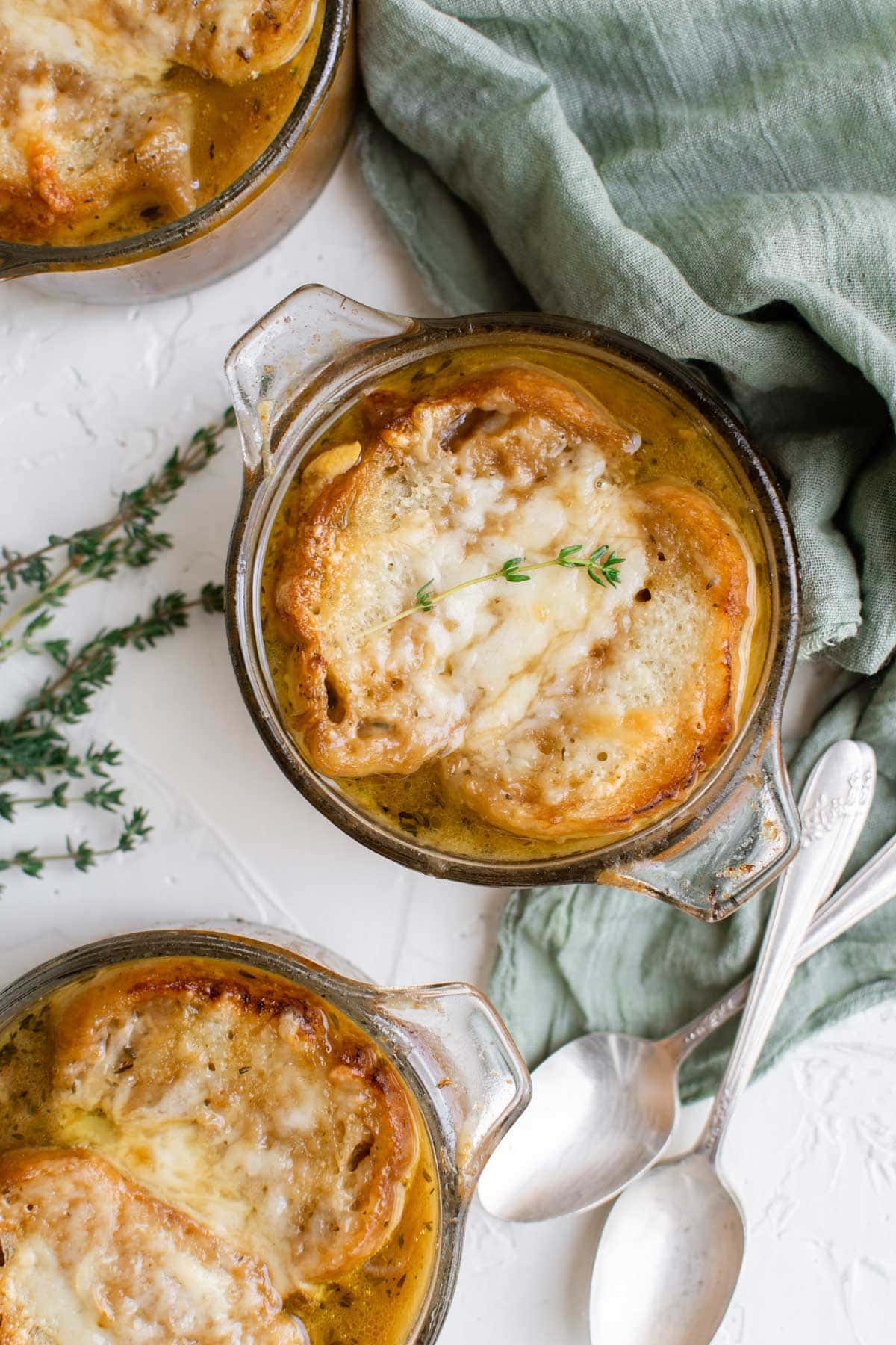 three clear glass soup bowls, tops of sou bowls with cheesy bread and onion soup, green napkin, spoons, thyme