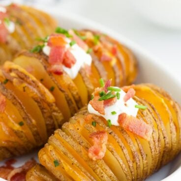 Hasselback Potatoes in a white serving dish with sour cream, bacon and fresh chives on top.