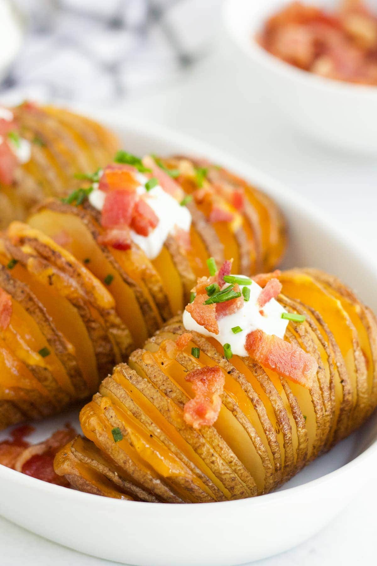 Hasselback Potatoes in a white serving dish with sour cream, bacon and fresh chives on top. 