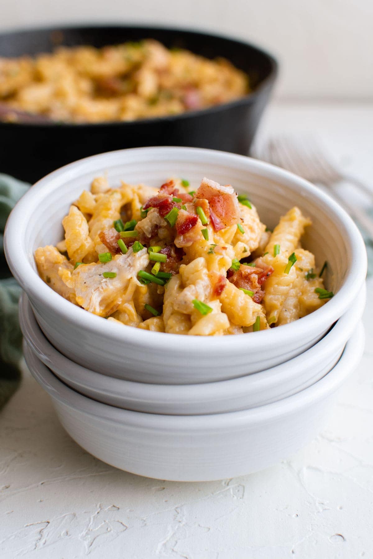 white bowls, stacked, with cheesy pasta and bacon, chives, cast iron skillet with pasta in the background