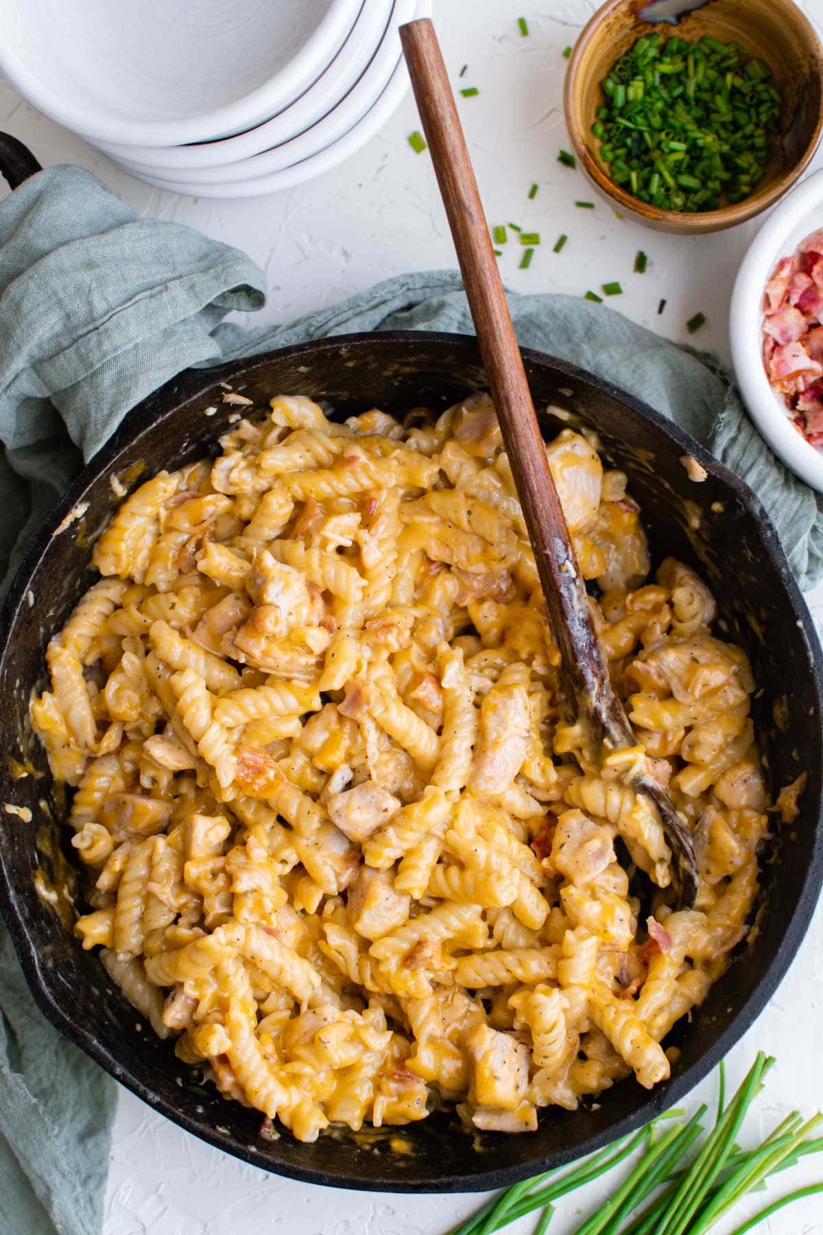 cast iron skillet with cheesy pasta with chicken and bacon, bowl of bacon, bowl of chives, empty white bowls, wooden spoon