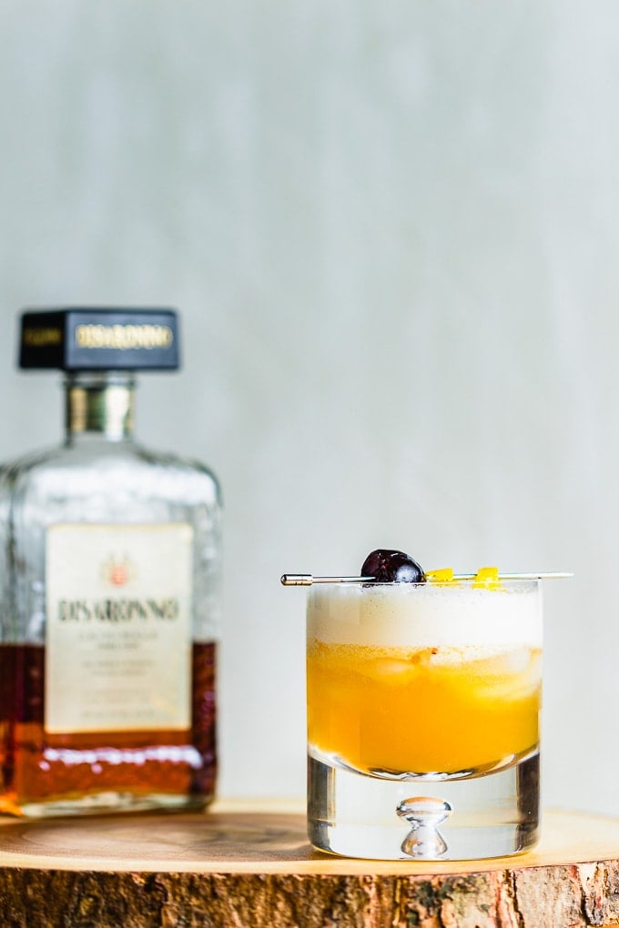 Rocks glass with amaretto sour garnished with cherry and lemon twist and a bottle of Amaretto.