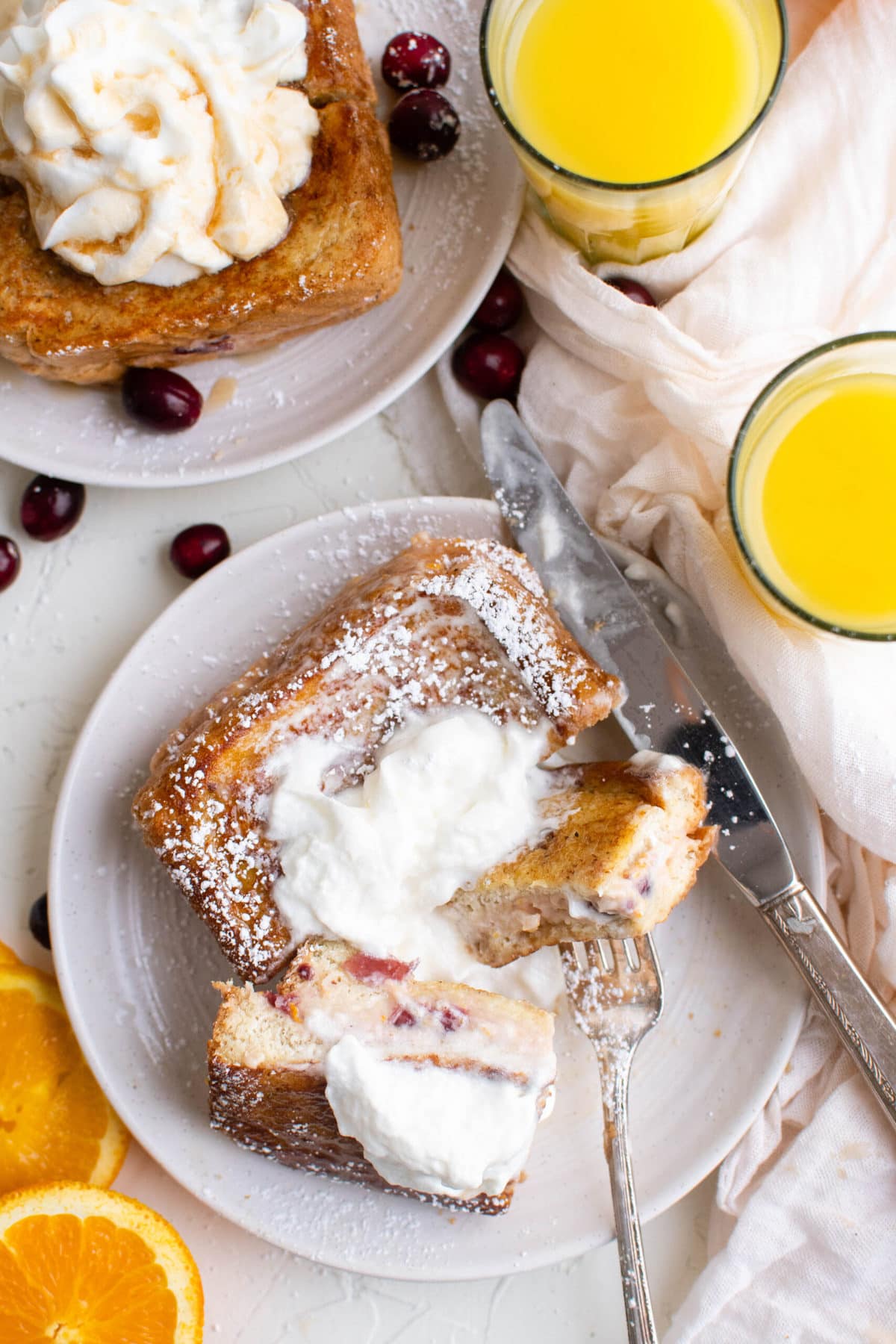 two white plates, cranberry cream cheese stuffed french toast sandwiches, fork, knife, glasses of orange uice, orange slices, cranberries