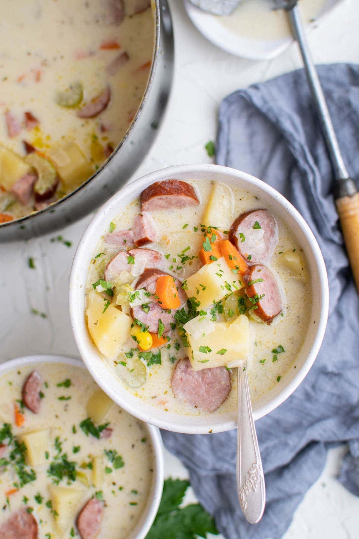 white bowls with sausage and potato chowder, spoons, blue napkin , a soup pot