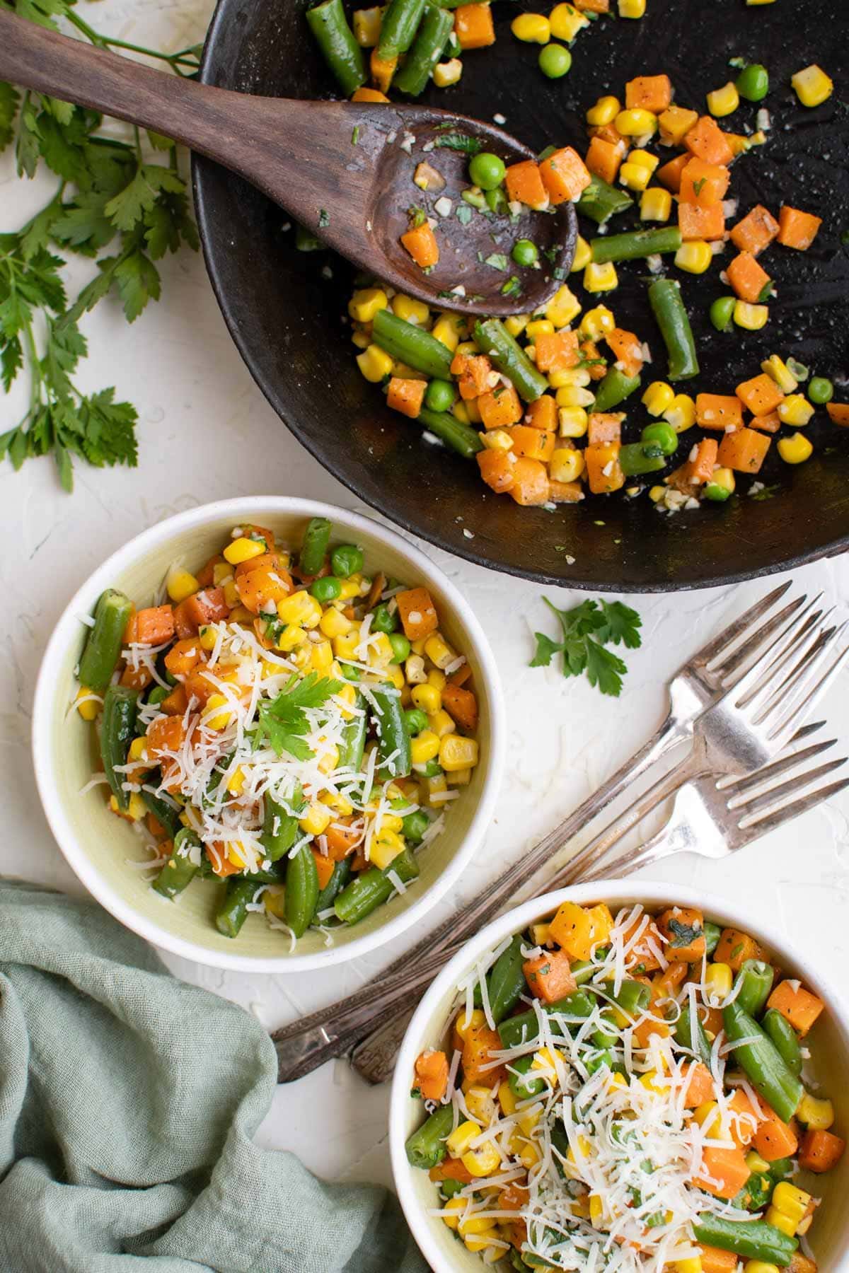 skillet with garlic butter vegetables, two white plates with veggies and parmesan cheese, wooden spoon, parsley sprigs, 3 forks, green napkin