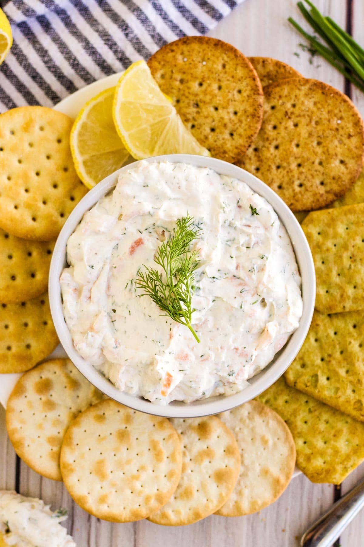 crackers, white dish filled with smoked salmon dip, dill, chives, blue and white striped napkin, lemon slices