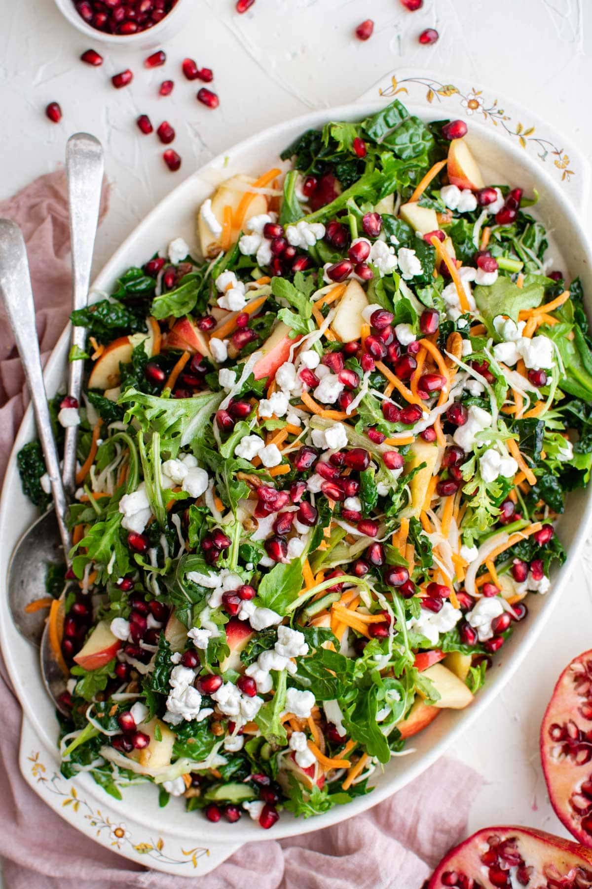 salad with pomegranates, apples, carrots, pecans and goat cheese on a white platter, metal spoons