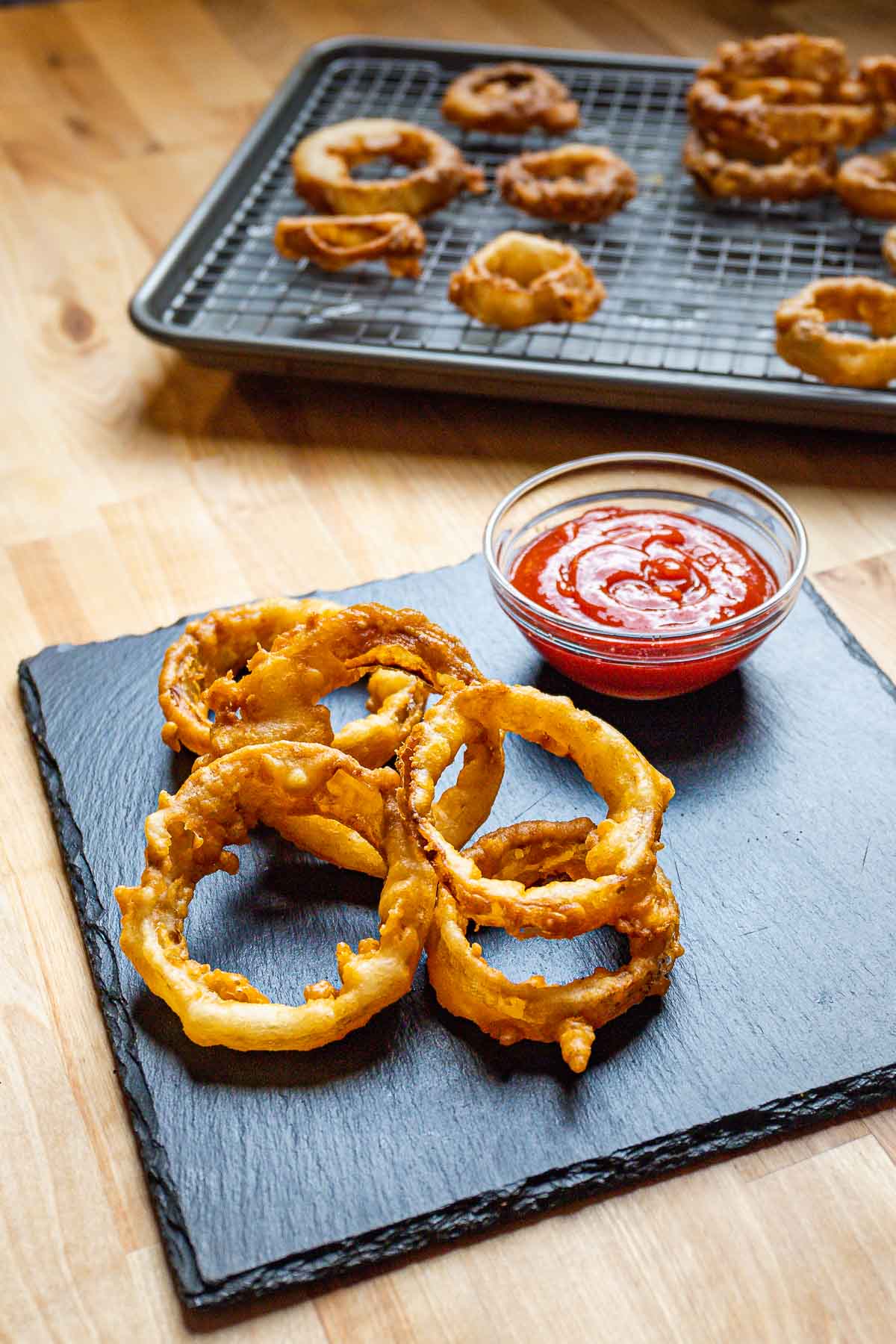 Onion rings and ketchup on table with sheet pan of extra onion rings in background.