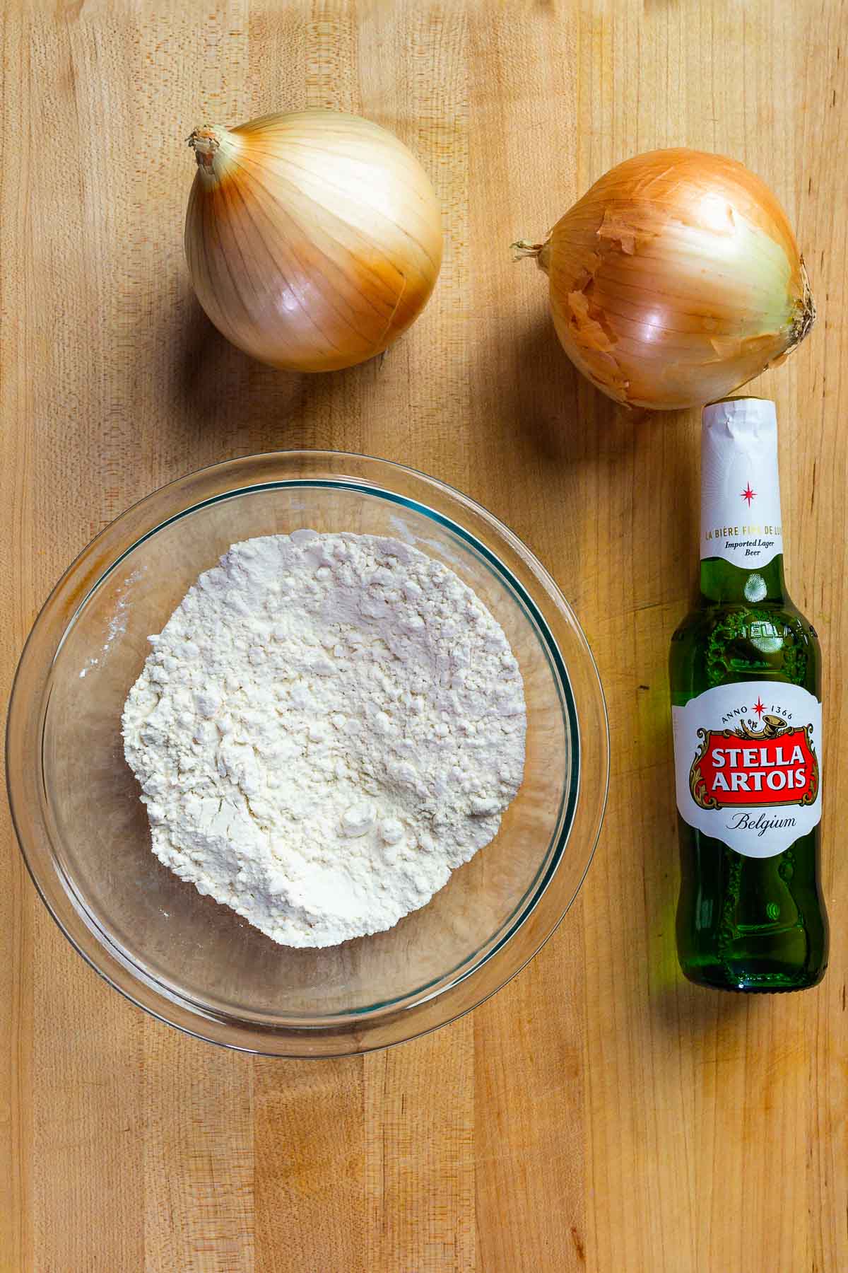 Two large onions, bowl of flour, and bottle of beer on wood cutting board.