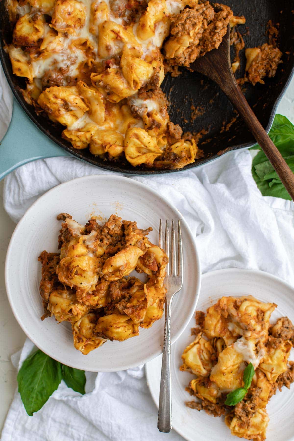 black skillet with tortellini casserole, white plates, fork, basil leave, white towel.