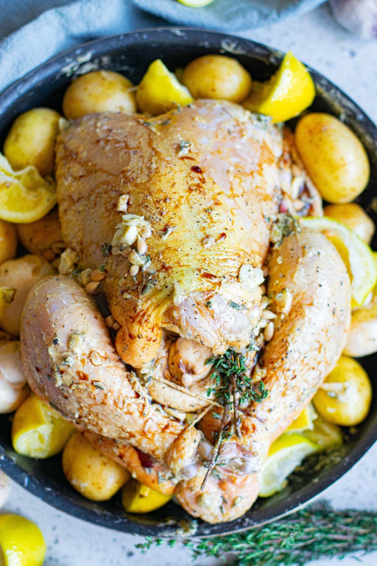 raw whole chicken with potatoes and lemon in a round pan, thyme sprigs, chicken has butter all over it.