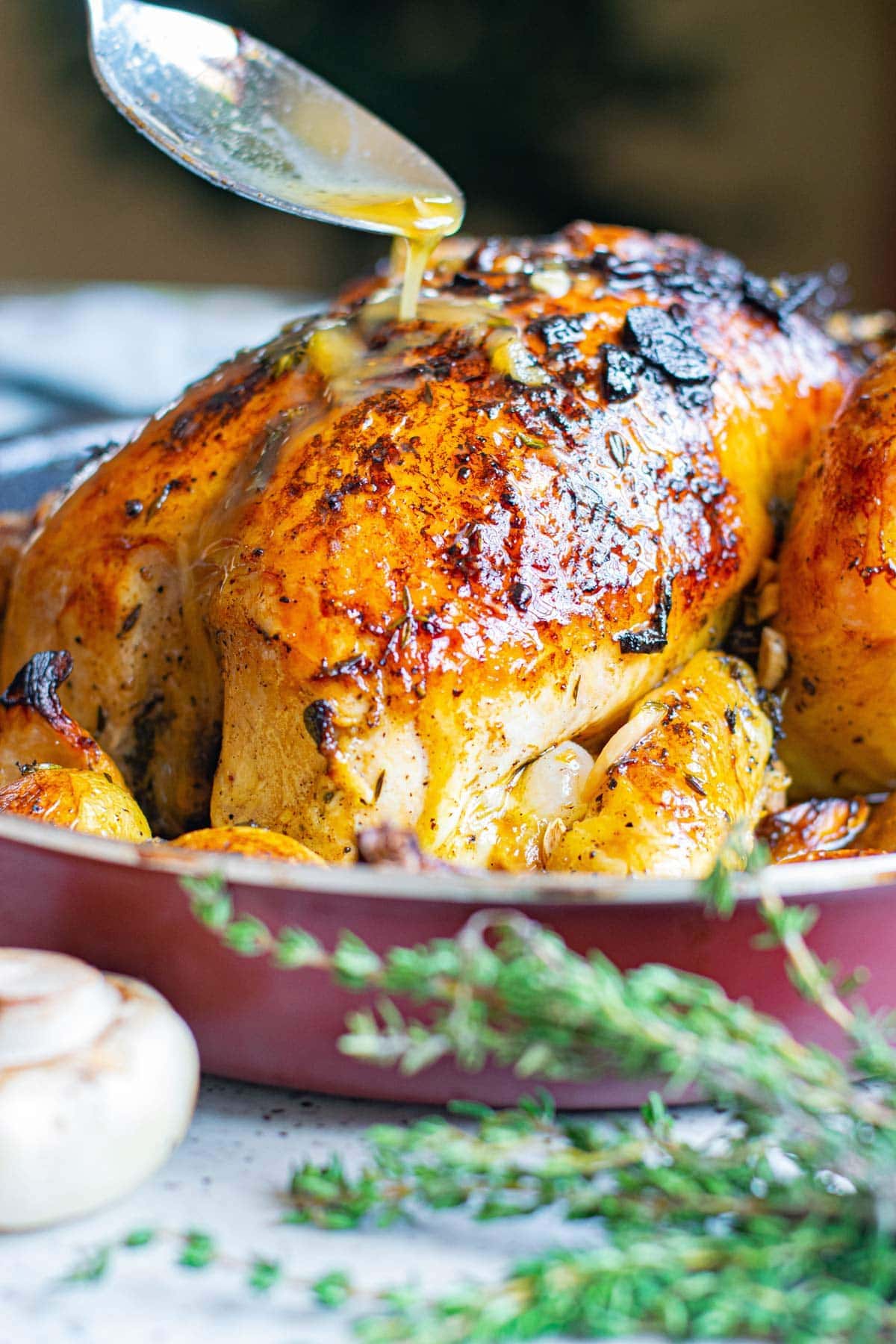 spoon basting juices over a whole chicken in a pan, thyme sprigs