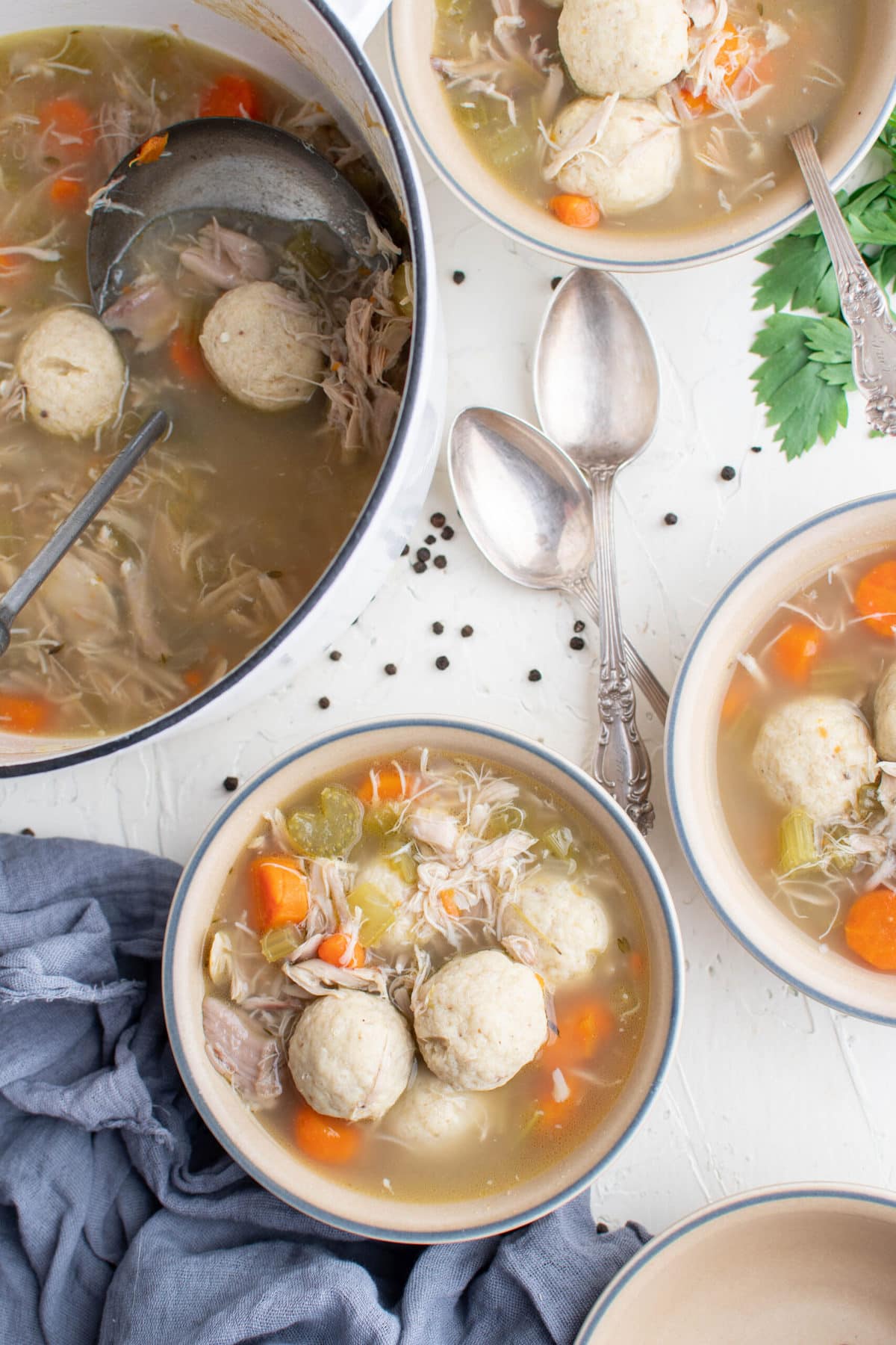 white bowl, matzo balls, pot of soup, spoons, parsley, blue napkin