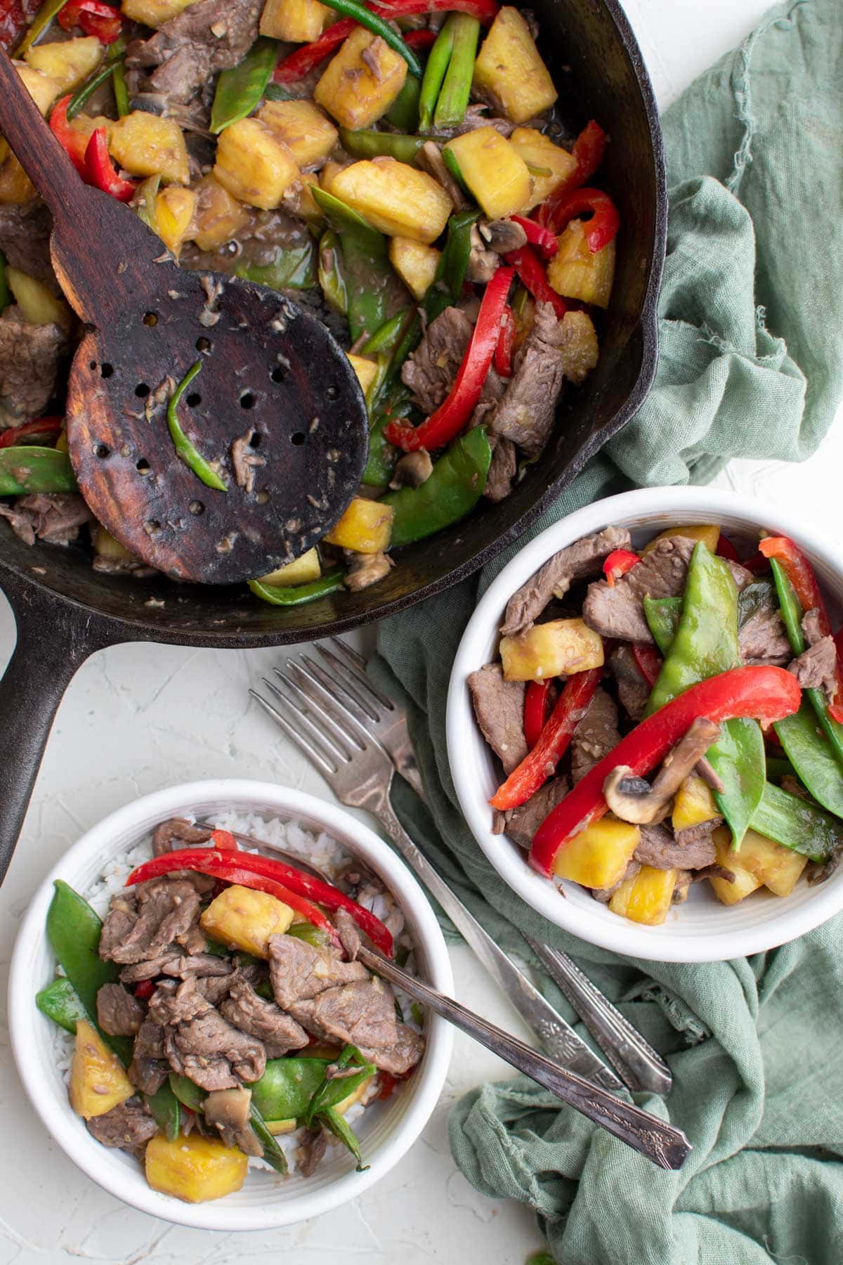 white bowls with beef, stir fry vegetables, green napkin, black skillet, wood spoon