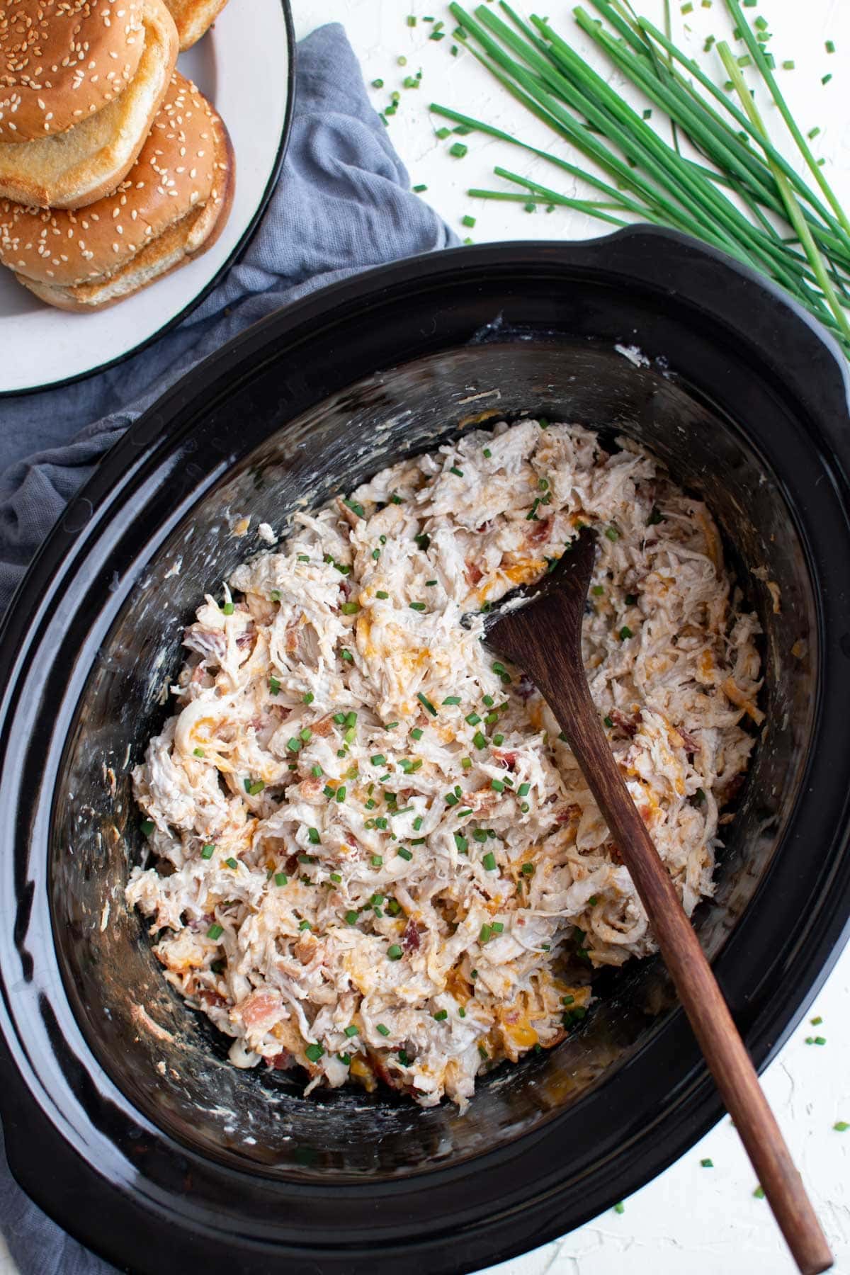 slow cooker pot with creamy shredded chicken, wooden spoon, stack of sesame seed buns, chives