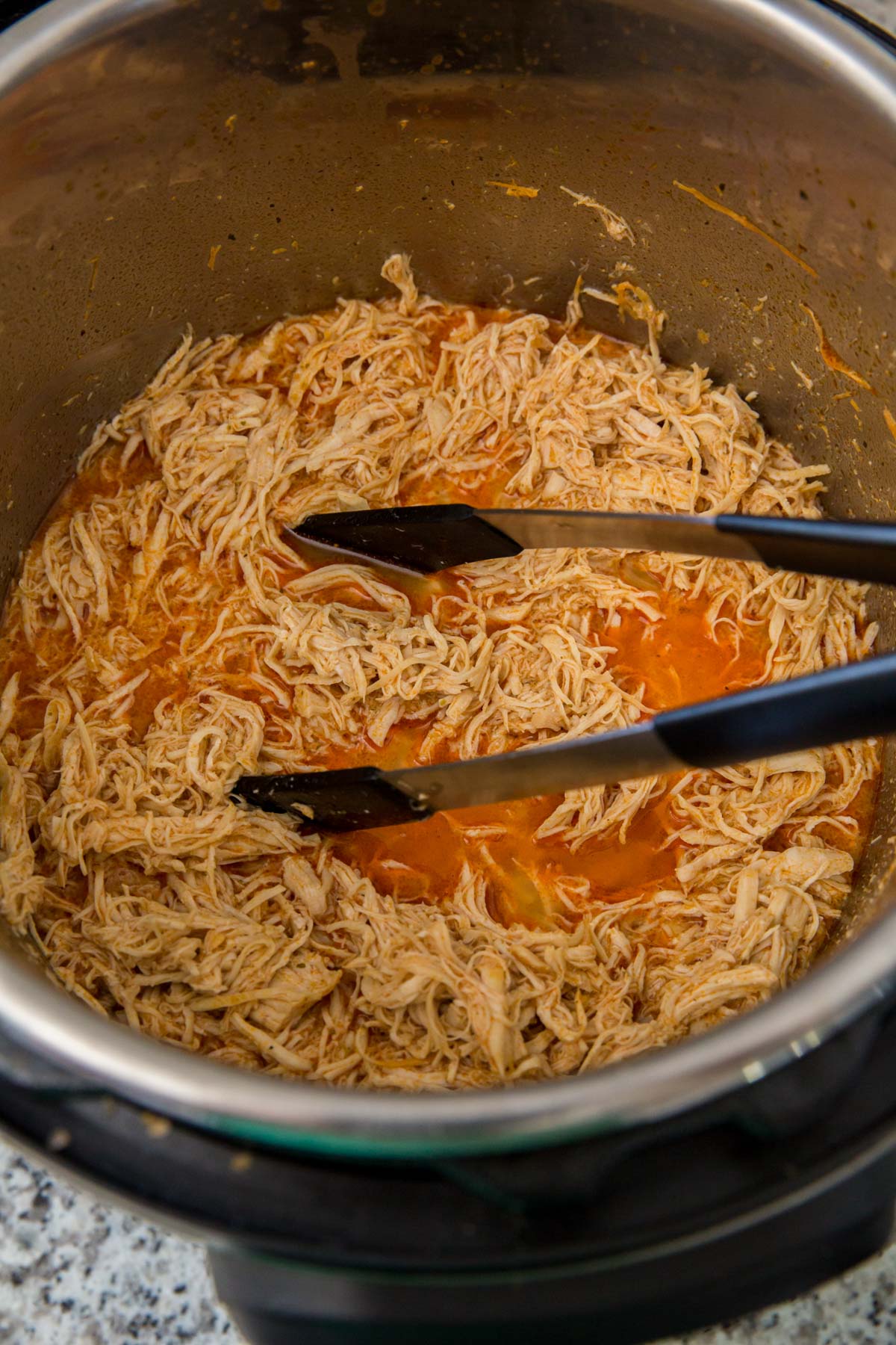 instant pot with shredded chicken and buffalo sauce, black and silver tongs