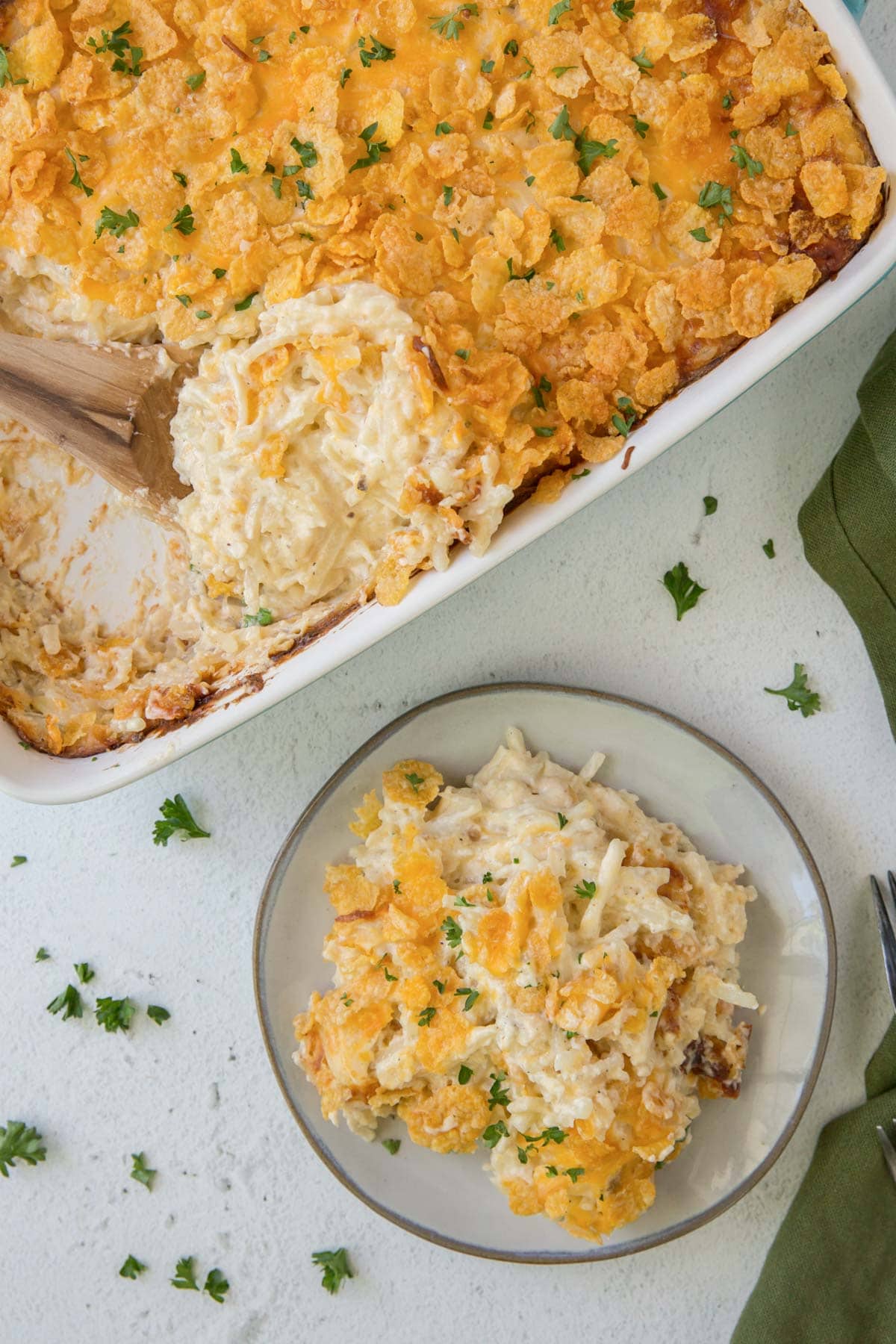 cracker barrel hashbrown casserole, casserole dish, corn flakes topping, wooden spoon, white plate, green napkin