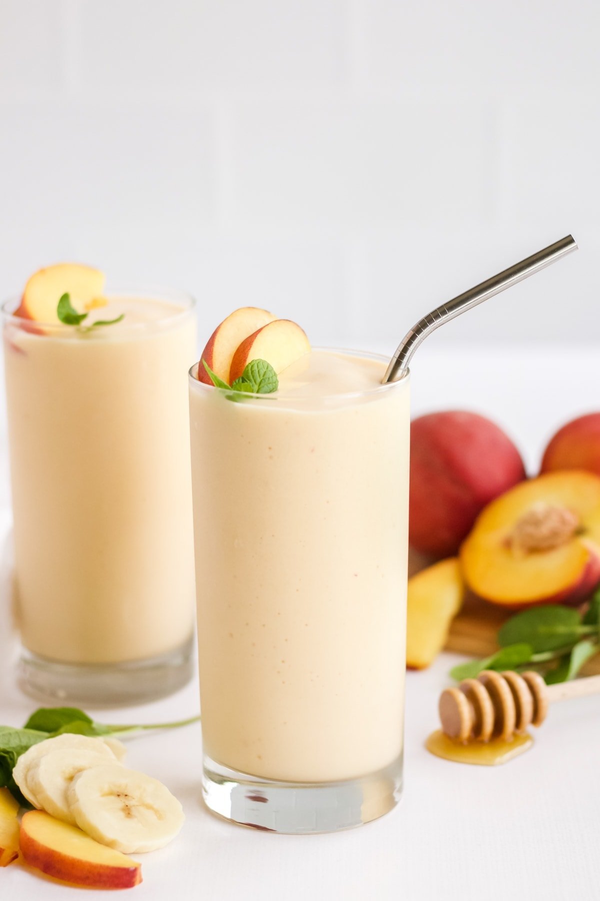 A picture of two peach banana smoothies with ingredients laid out around them and a white backdrop