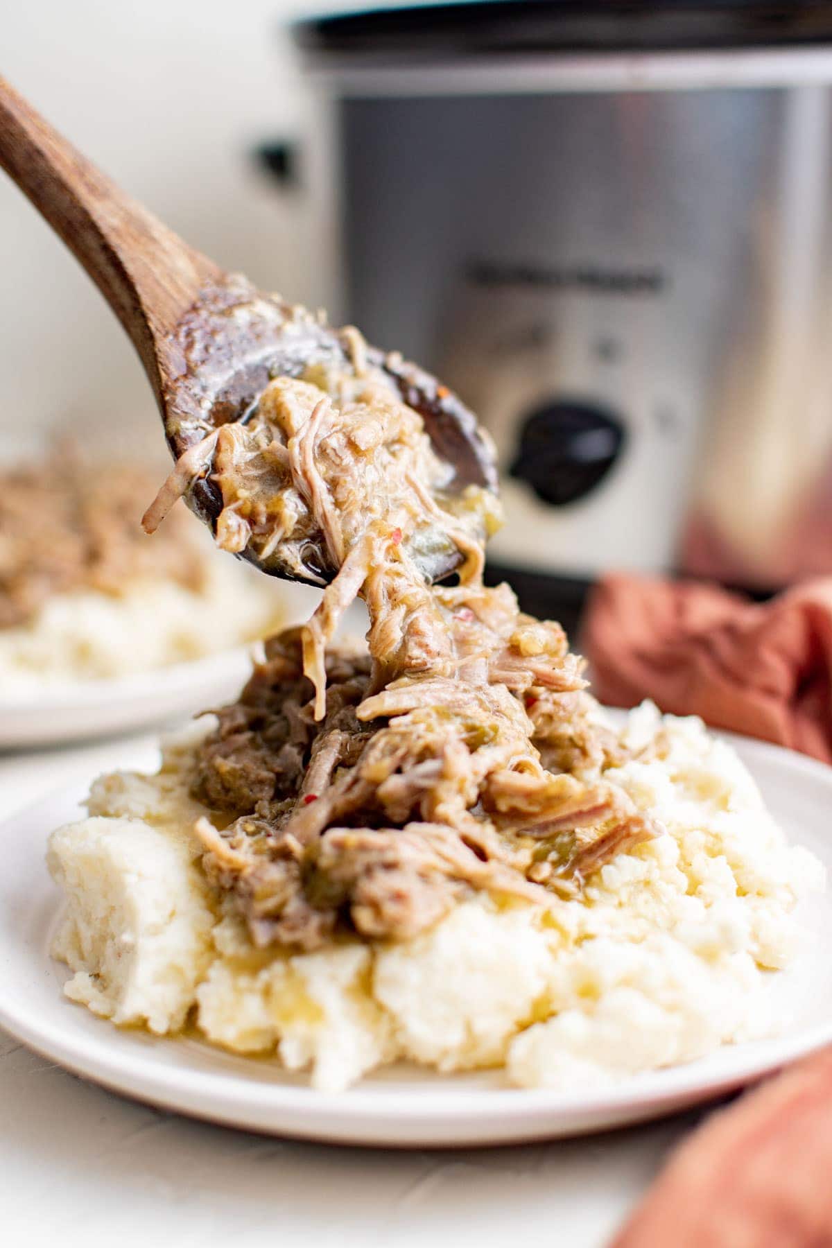 white plate, wooden spoon serving chile verde onto mashed potatoes, slow cooker