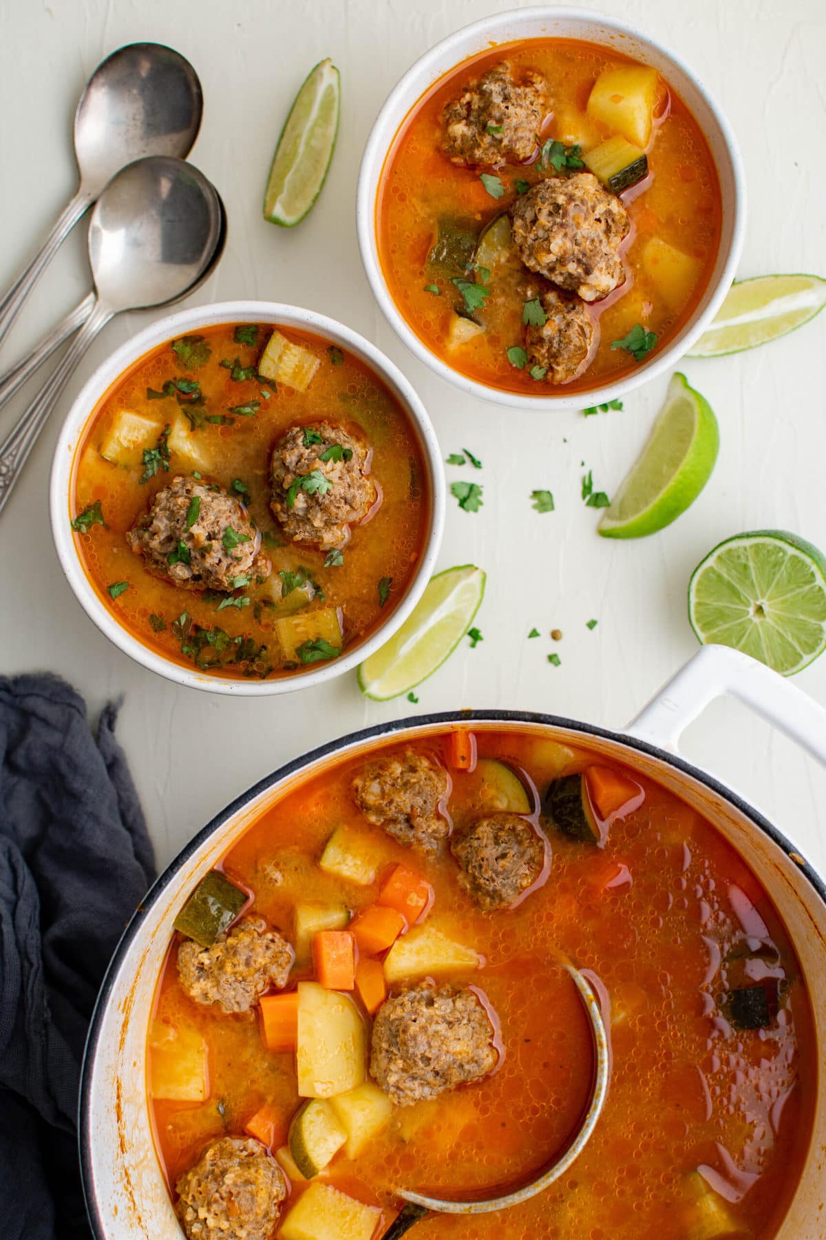 pot of mexican meatball soup, spoons, white bowls, limes