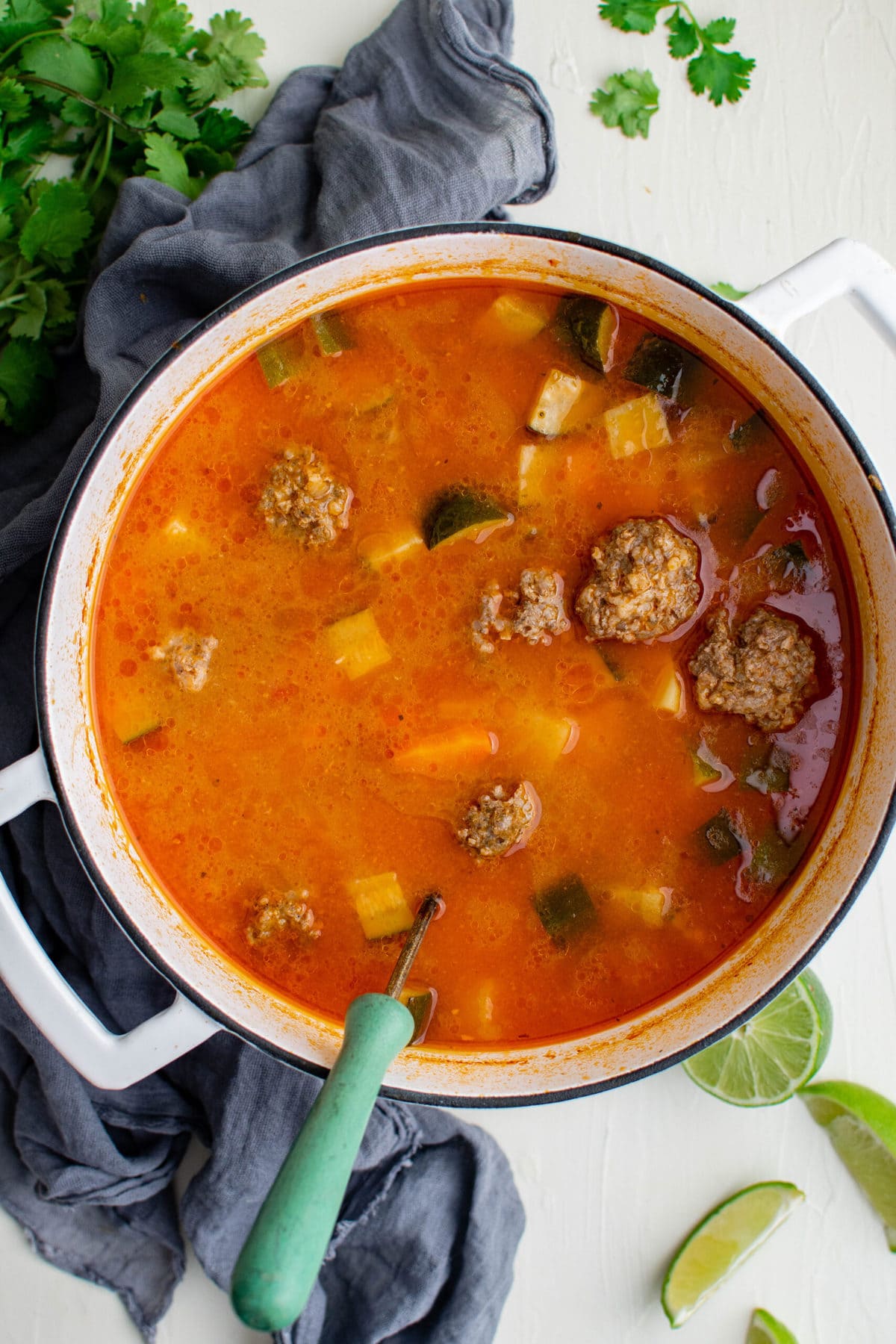 white soup pot, ladle, blue napkin, cilantro, limes, albondigas soup