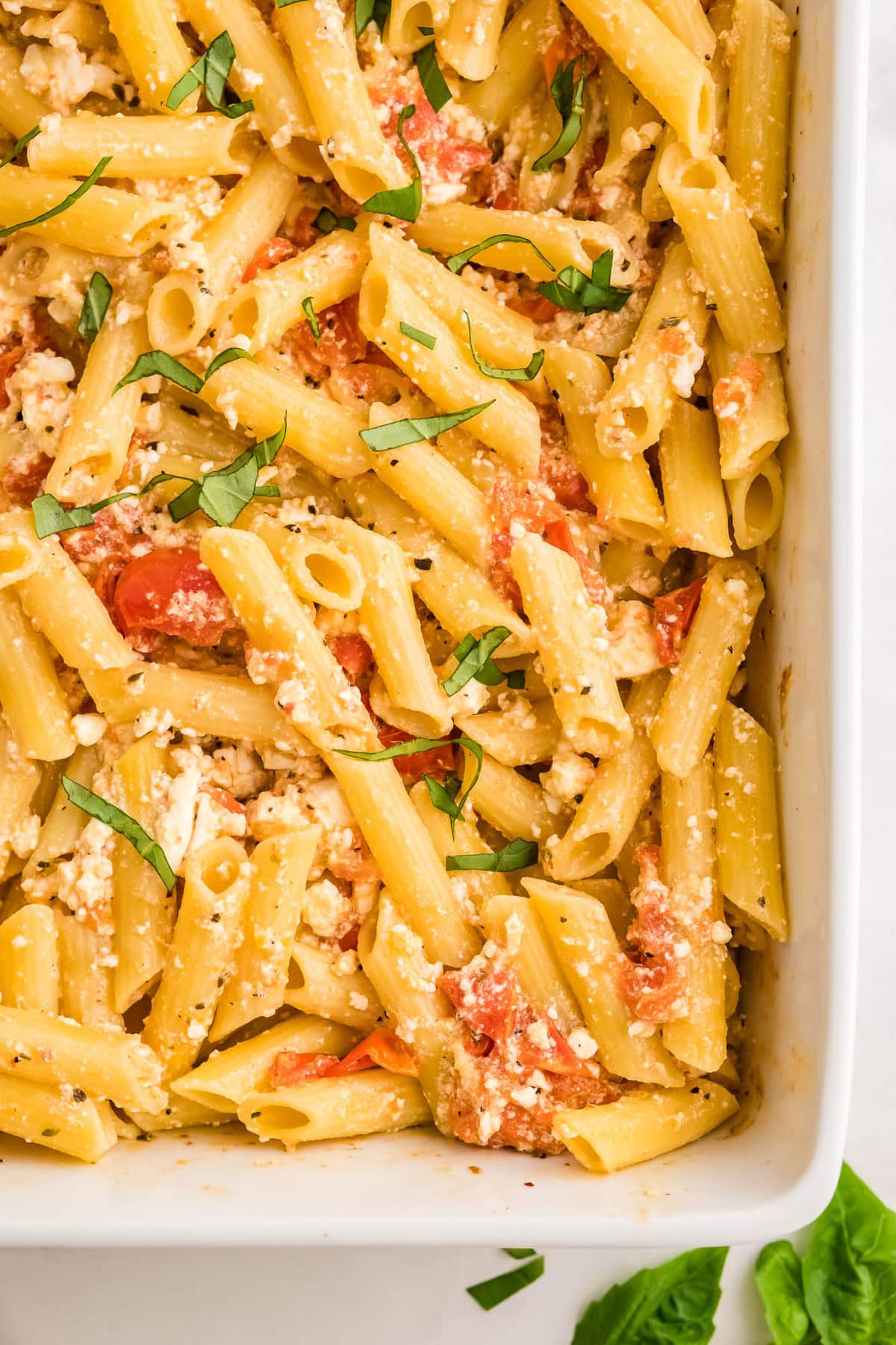 white baking dish, penne pasta, basil, tomatoes and feta