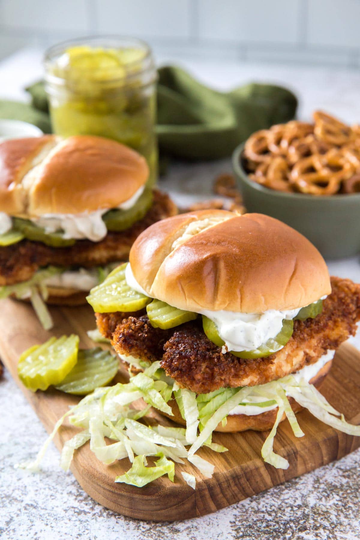 crispy chicken sandwiches on a wood board with lettuce and pickles, bowl of pretels