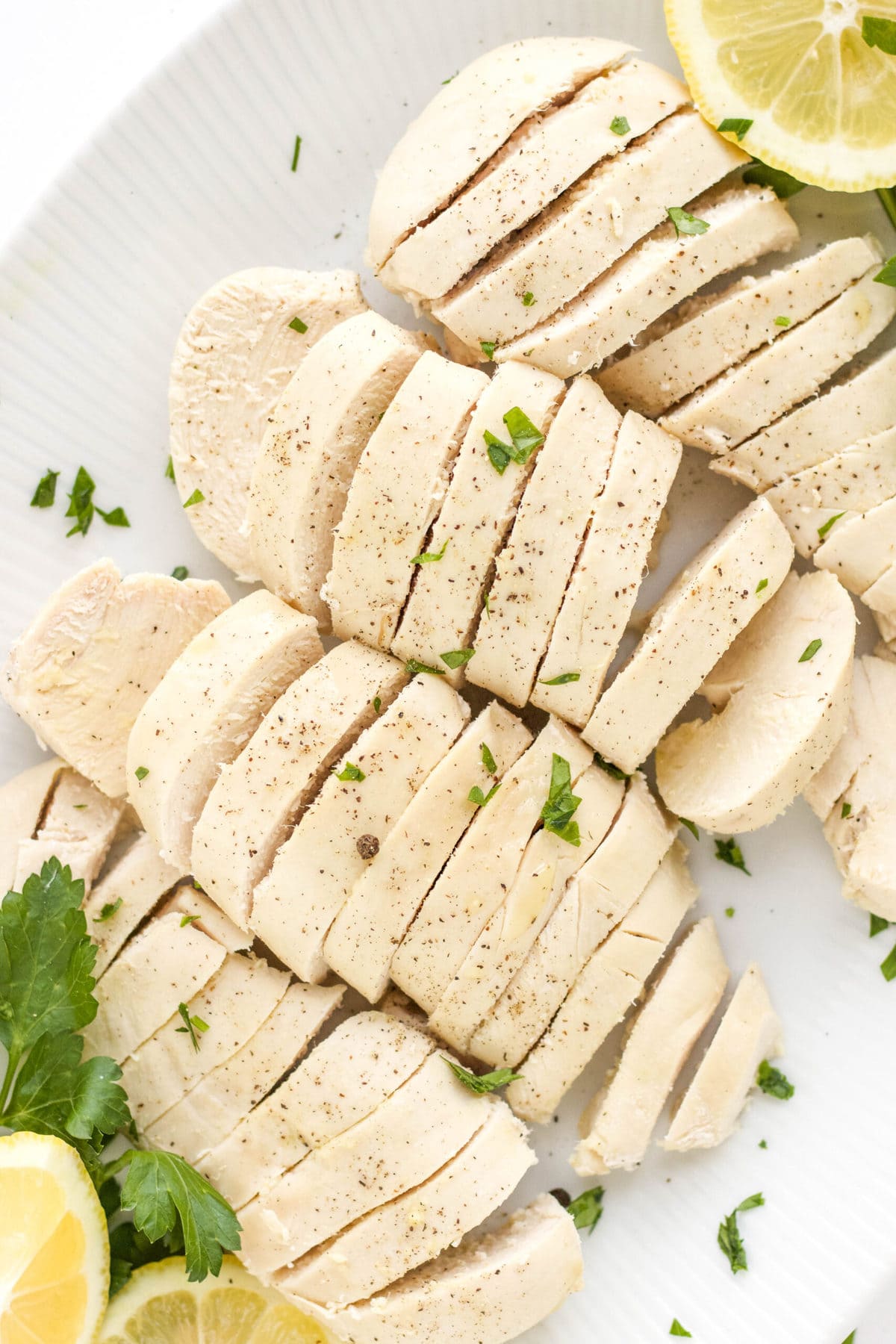 sliced poached chicken brerasts on a white platter with parsley and lemons