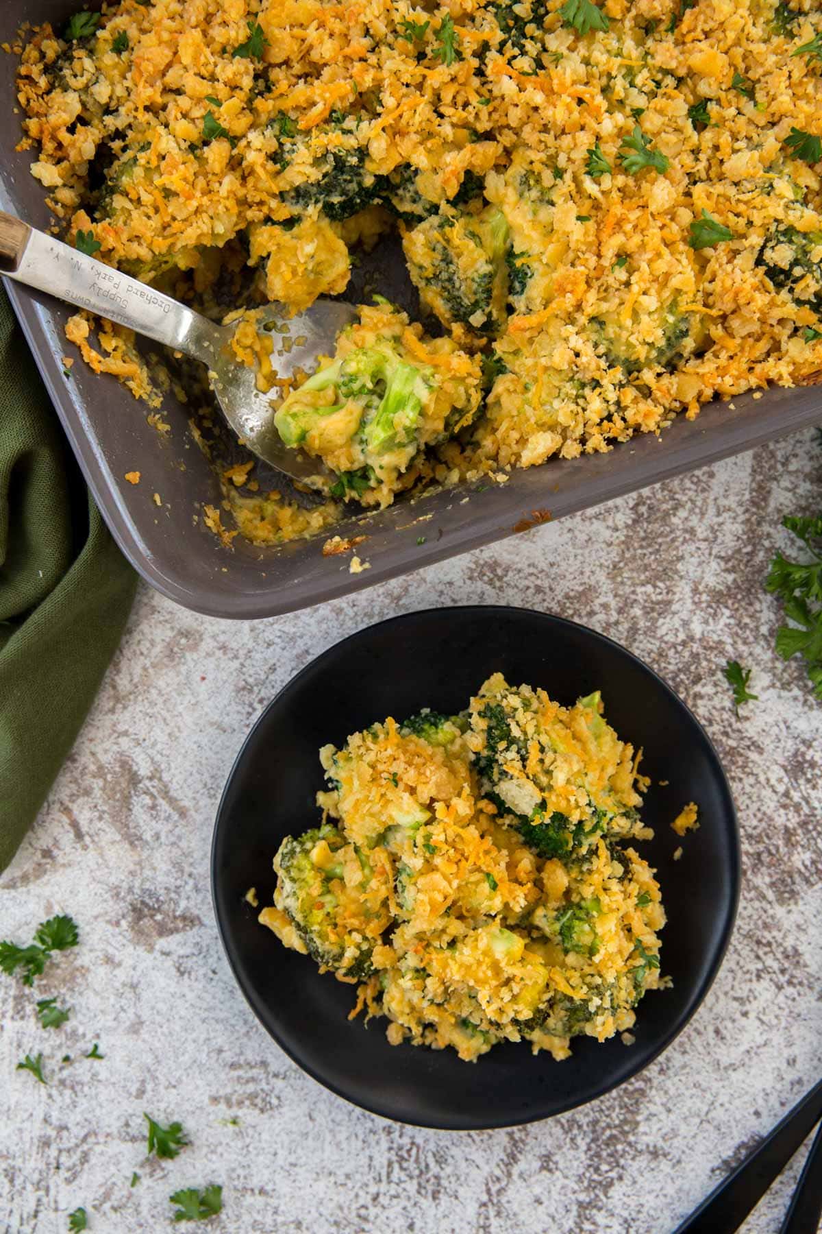 black plate, gray baking dish, broccoli, cheese sauce, crushed cracker topping, large serving spoon