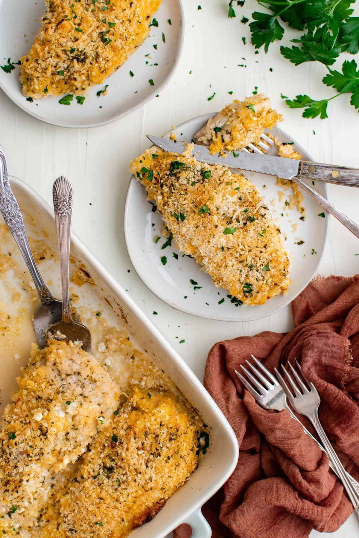 baking dish, chicken breasts with breadcrumb topping, pink napkin, forks, parsley, white plates