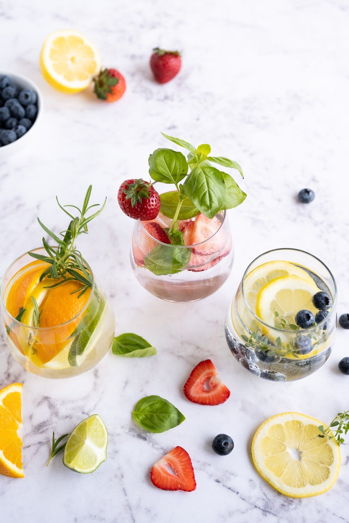 Three glasses with infused water in three glasses with strawberries, citrus fruit, fresh herbs, and blueberries.