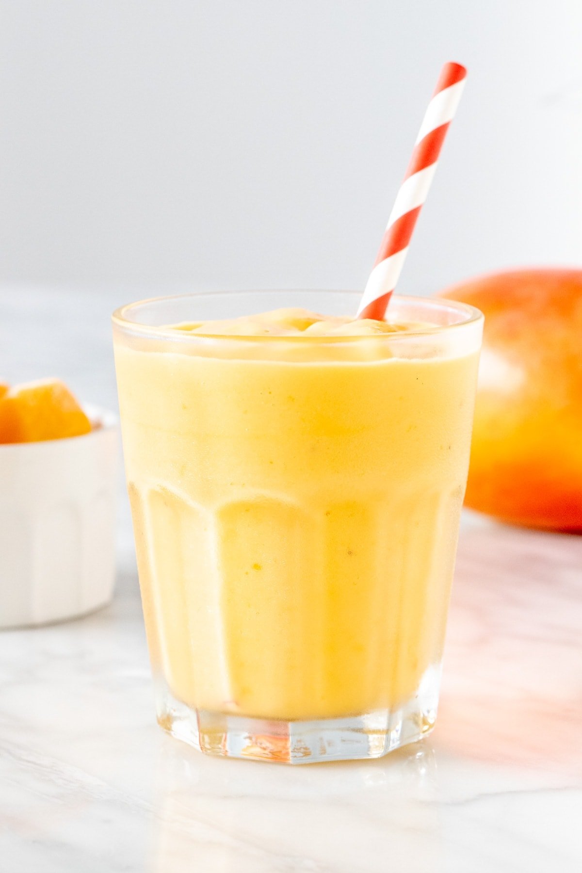 Glass of mango smoothie, red and white striped straw, whit bowl with cut mango, whole mangu