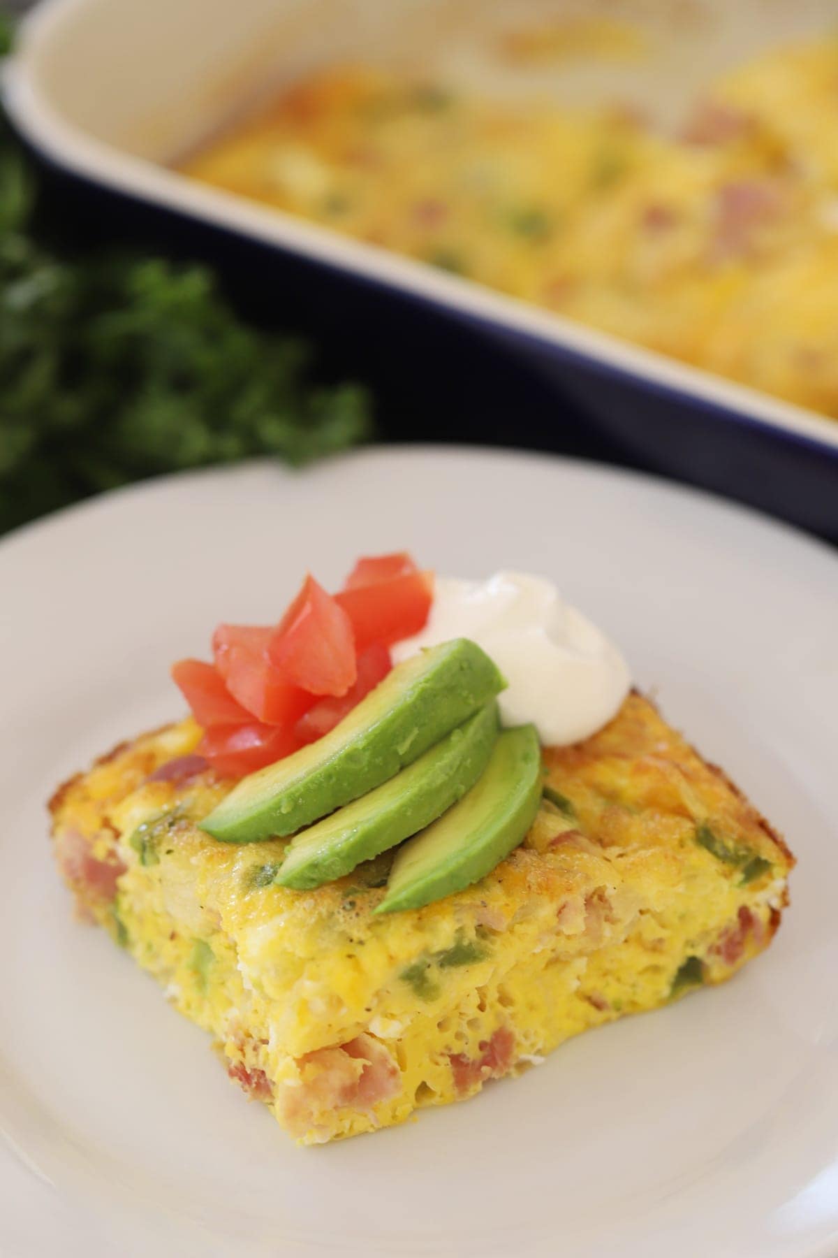 Baked Denver Omelet on a white plate, covered in sour cream, diced tomatoes and sliced avocado