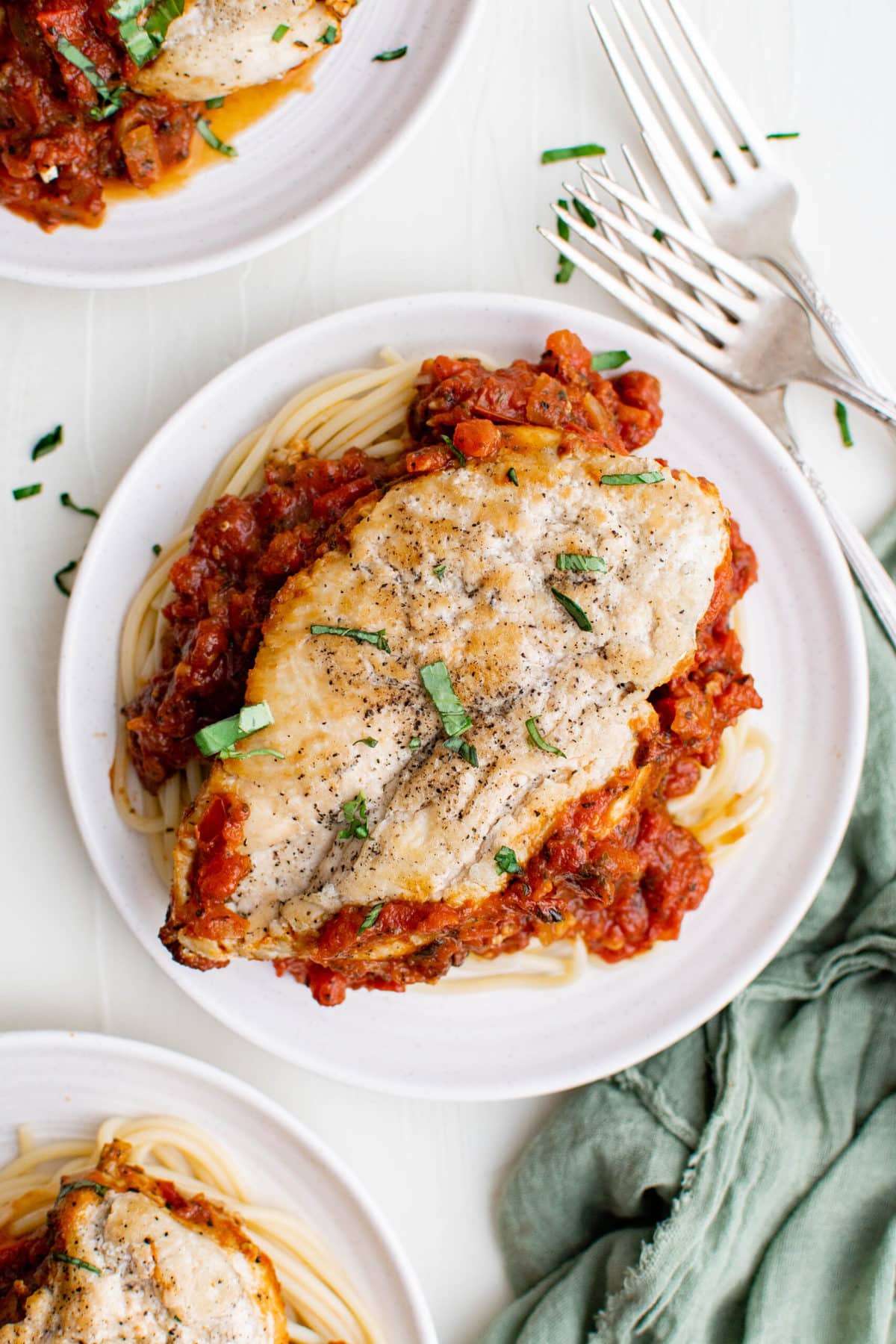 chicken breast over tomato pomodoro sauce and pasta on white plates, fork, knife, green napkin