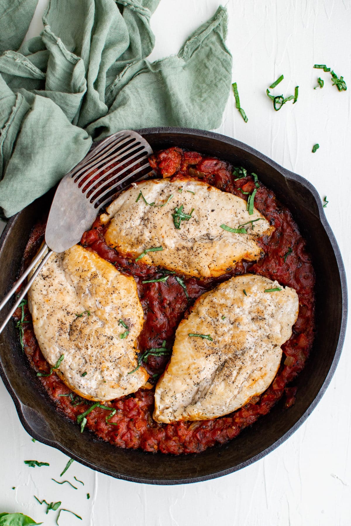 3 chicken breasts with tomato sauce in black skillet, metal spatula, green napkin