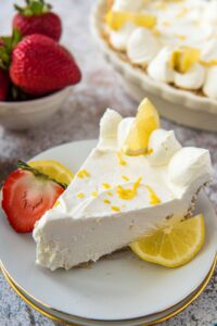 A slice of lemon cheesecake on a white plate with strawberries and lemon on the side