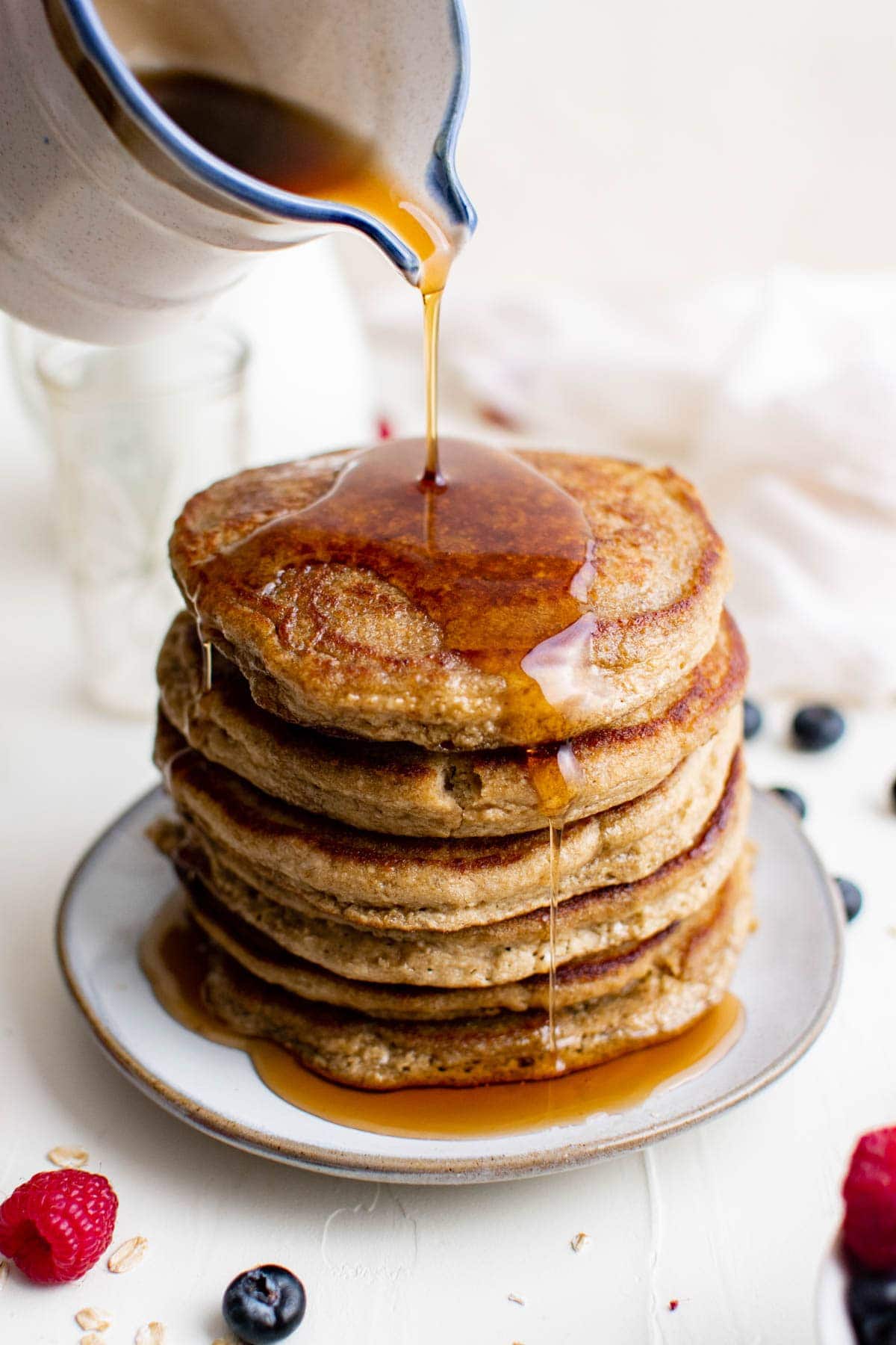 stack of pancakes on a white plate, syrup pouring fron a white pitcher, berries