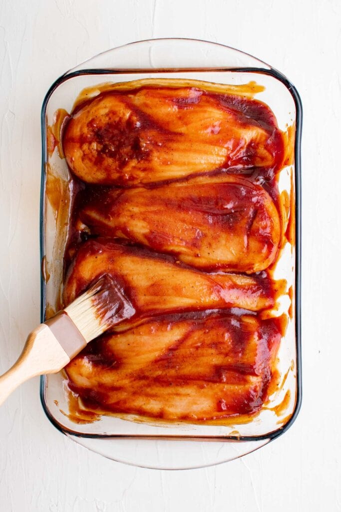 chicken breasts in a baking dish with barbecue sauce and a basting brush