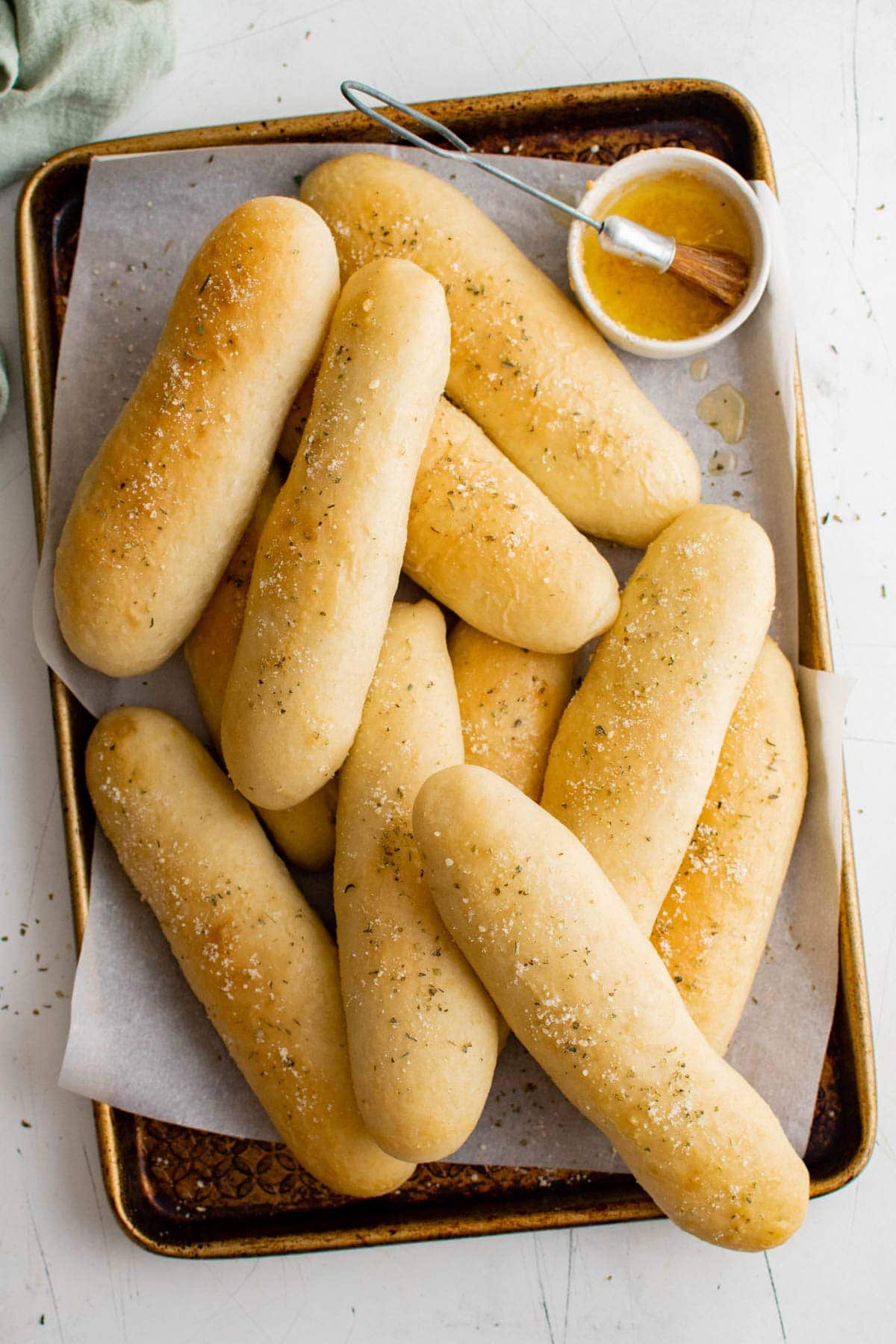 homemade garlic breadsticks piled on a baking sheet with garlic butter in a dish and a pastry brush