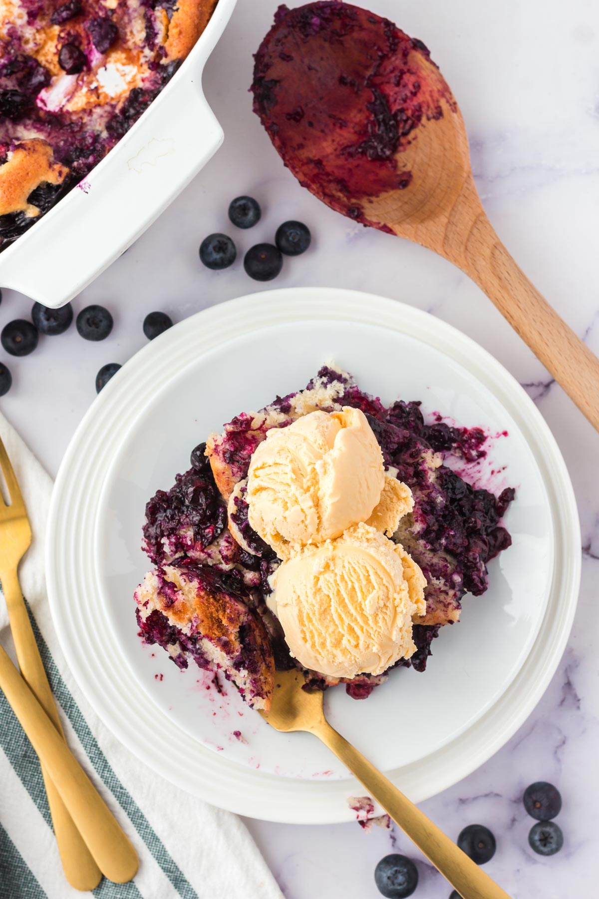 A white plate with blueberry cobbler and vanilla ice crea, a gold fork and a wooden spoon