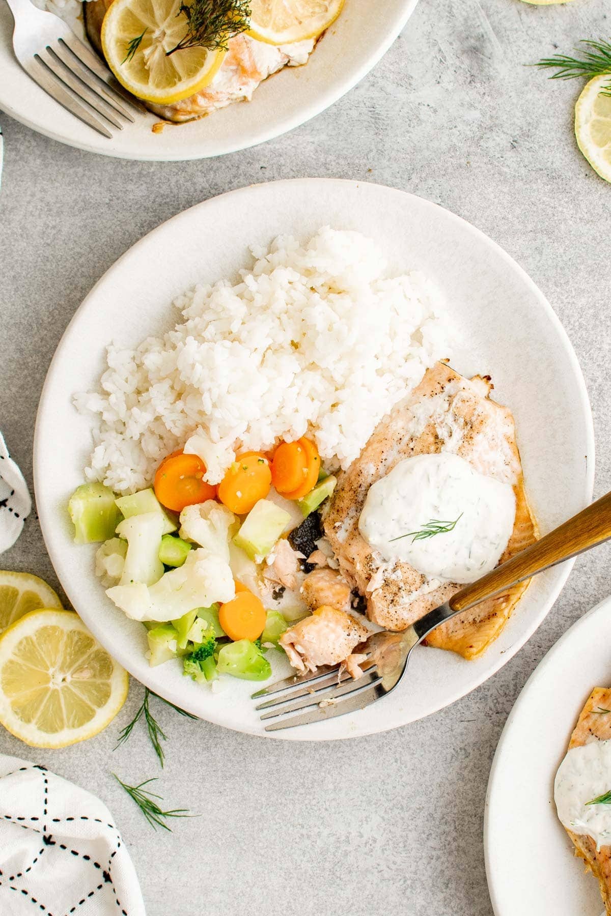 A piece of salmon with creamy dill sauce, rice and vegetables on a white plate, with a fork.