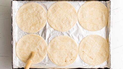 6 corn tortillas on a foil lined baking sheet with a pastry brush.