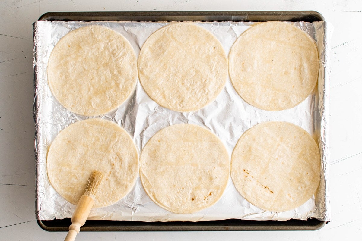 6 corn tortillas on a foil lined baking sheet with a pastry brush.