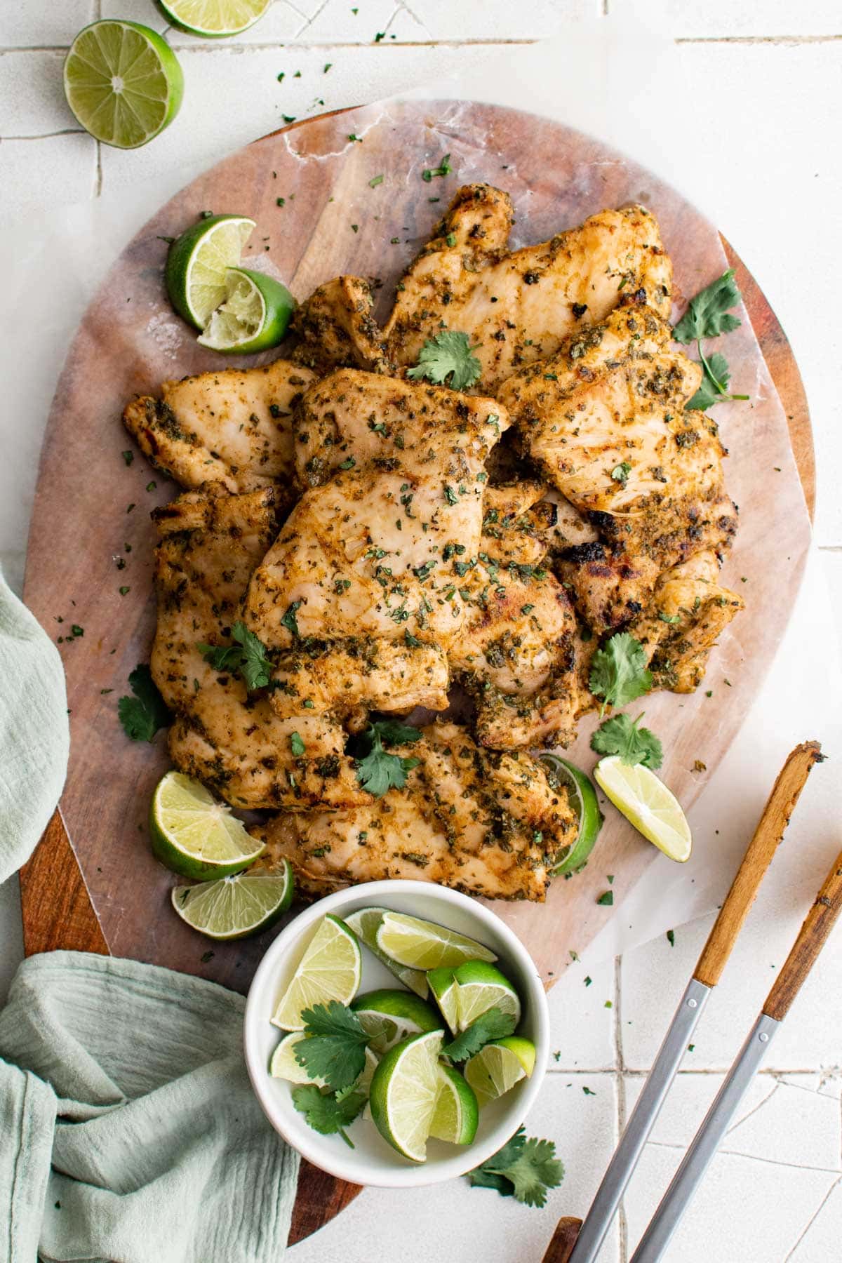 Wood platter with cilantro lime marinated chicken thighs, limes, tongs.