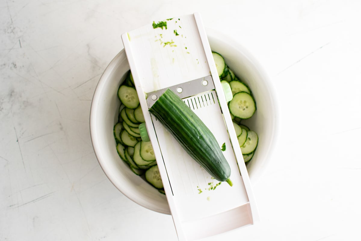 Cucumber resting on a madolin with sliced cucumbers below in a bowl.