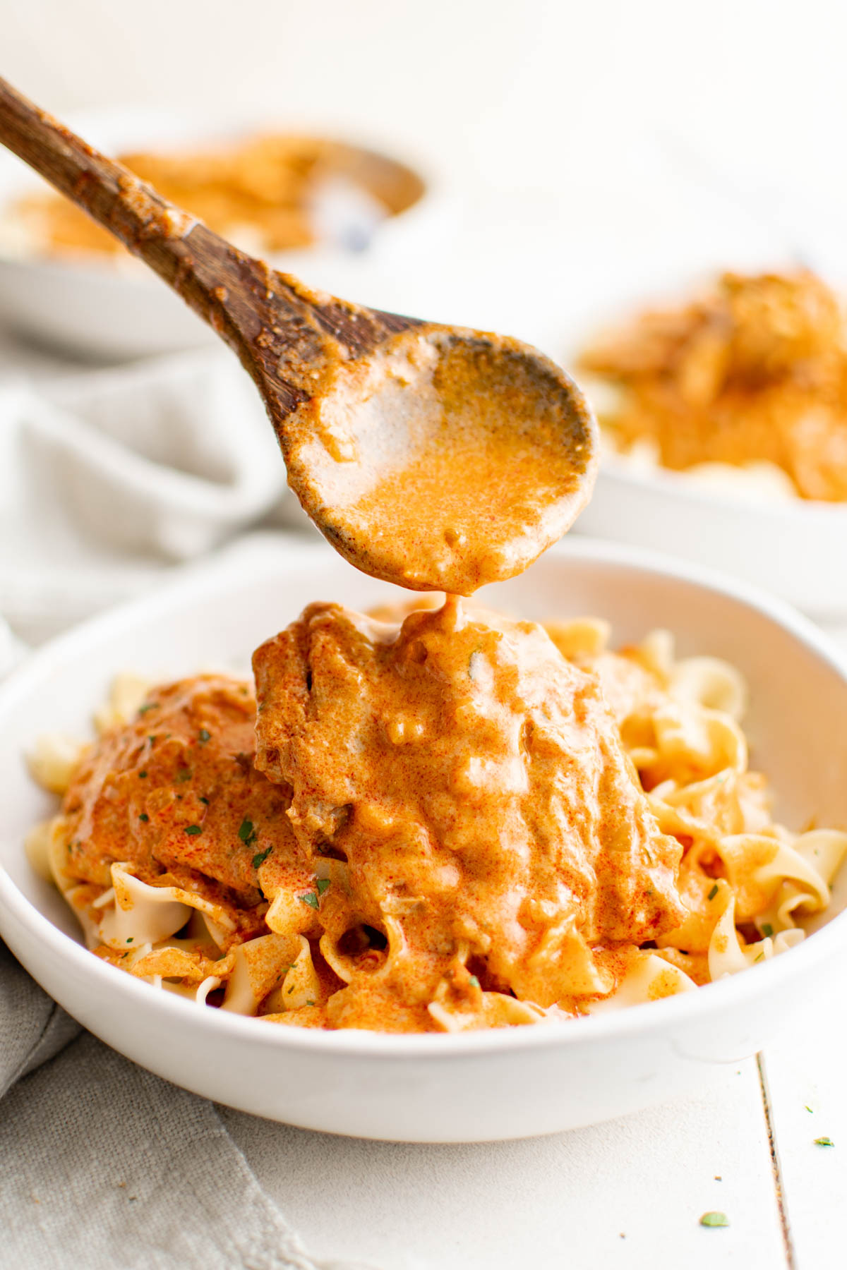 White bowl with egg noodles and chicken in paprikash sauce with wooden spoon pouring sauce over the top.