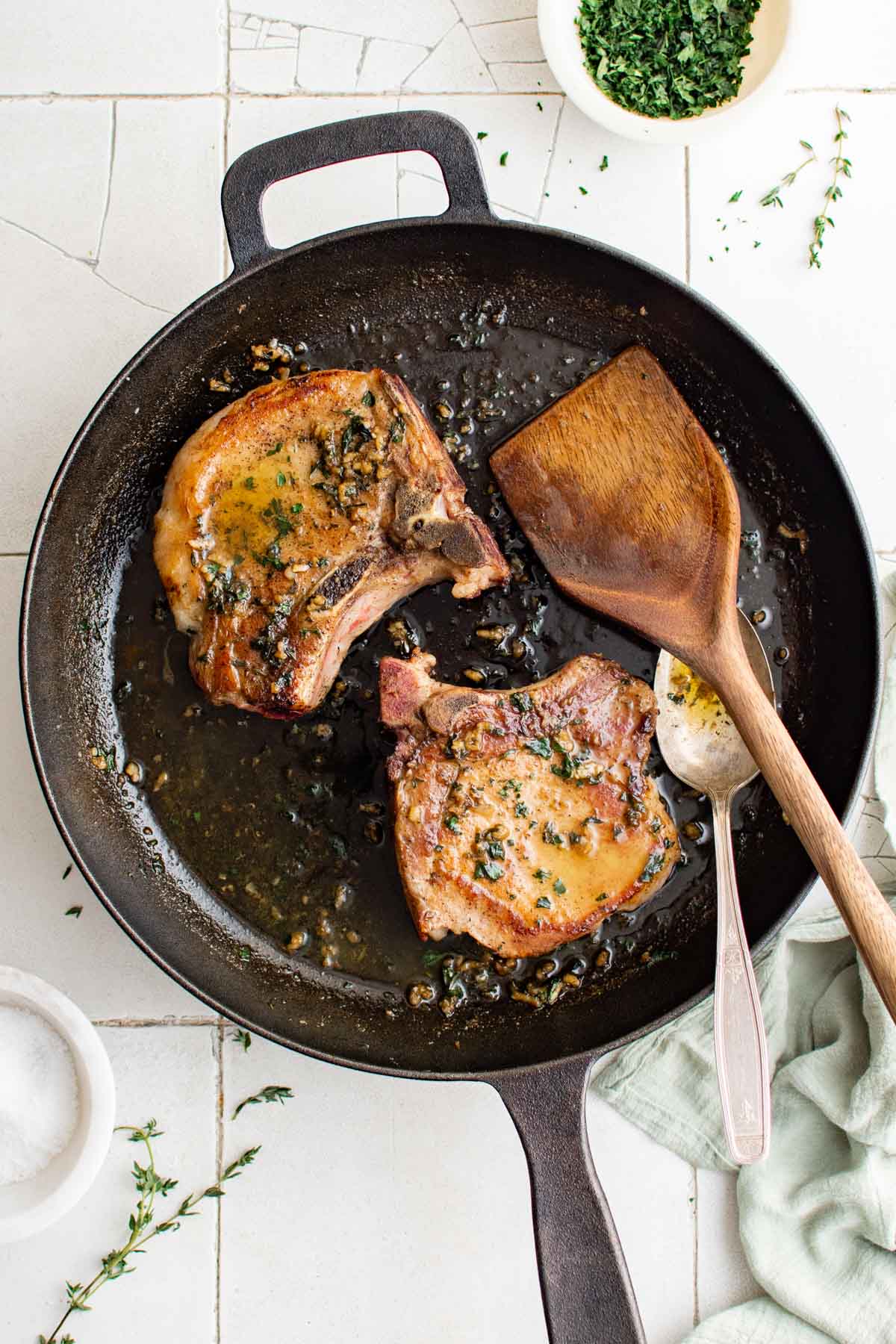 Two bone-in pork chops in a cast iron skillet with a wooden spatula.